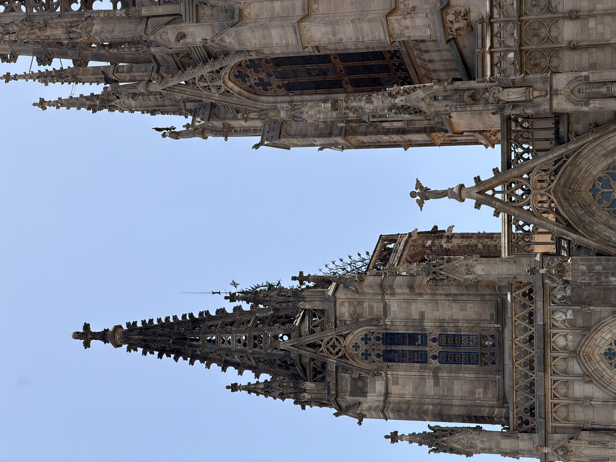 The image shows a close-up, low-angle view of the Gothic architecture of the Barcelona Cathedral, also known as the Cathedral of the Holy Cross and Saint Eulalia, located in the Gothic Quarter of Barcelona, Spain. The focus is on the intricate stonework of the cathedral's spires and facades against a clear, pale blue sky. Several tall, elaborately decorated spires dominate the frame, adorned with crosses, pinnacles, and ornamental tracery. Gargoyles are visible on the lower parts of the structures. A statue of an angel with outstretched wings stands atop one of the spires. The stonework is weathered, suggesting age. A large, multi-paned stained-glass window with a trefoil pattern is visible on one of the towers. The scene is devoid of people. The lighting suggests a bright, sunny day.