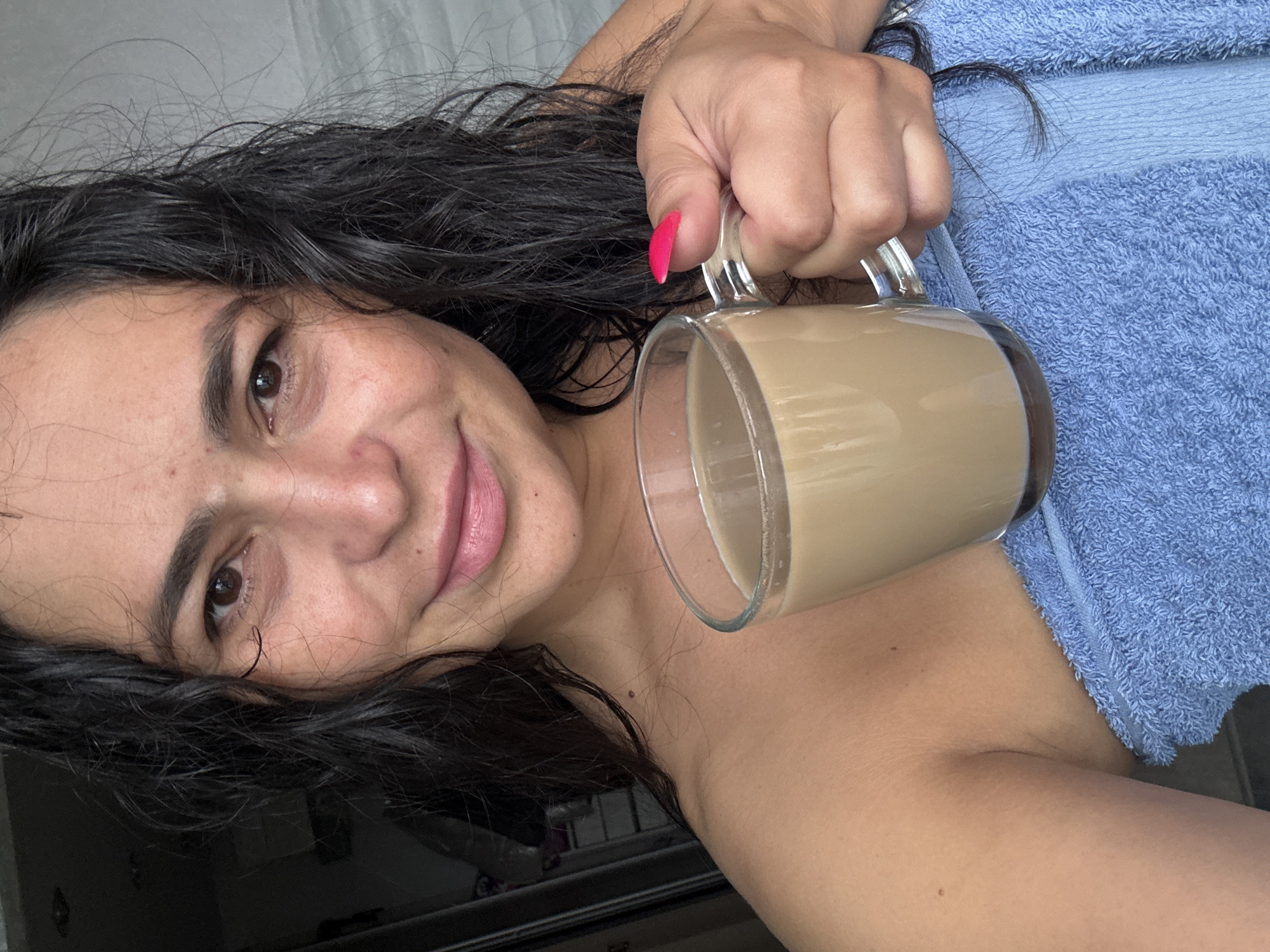 A person with dark hair, holding a clear mug containing a light brown liquid, is oriented at an angle in the image. The person has pink lipstick and a pink fingernail on the hand holding the mug. A blue towel is draped behind the person's arm. The environment appears to be indoors. There is no information available in this image to associate it with the city of Kauchuk Stantsiya, Russia.