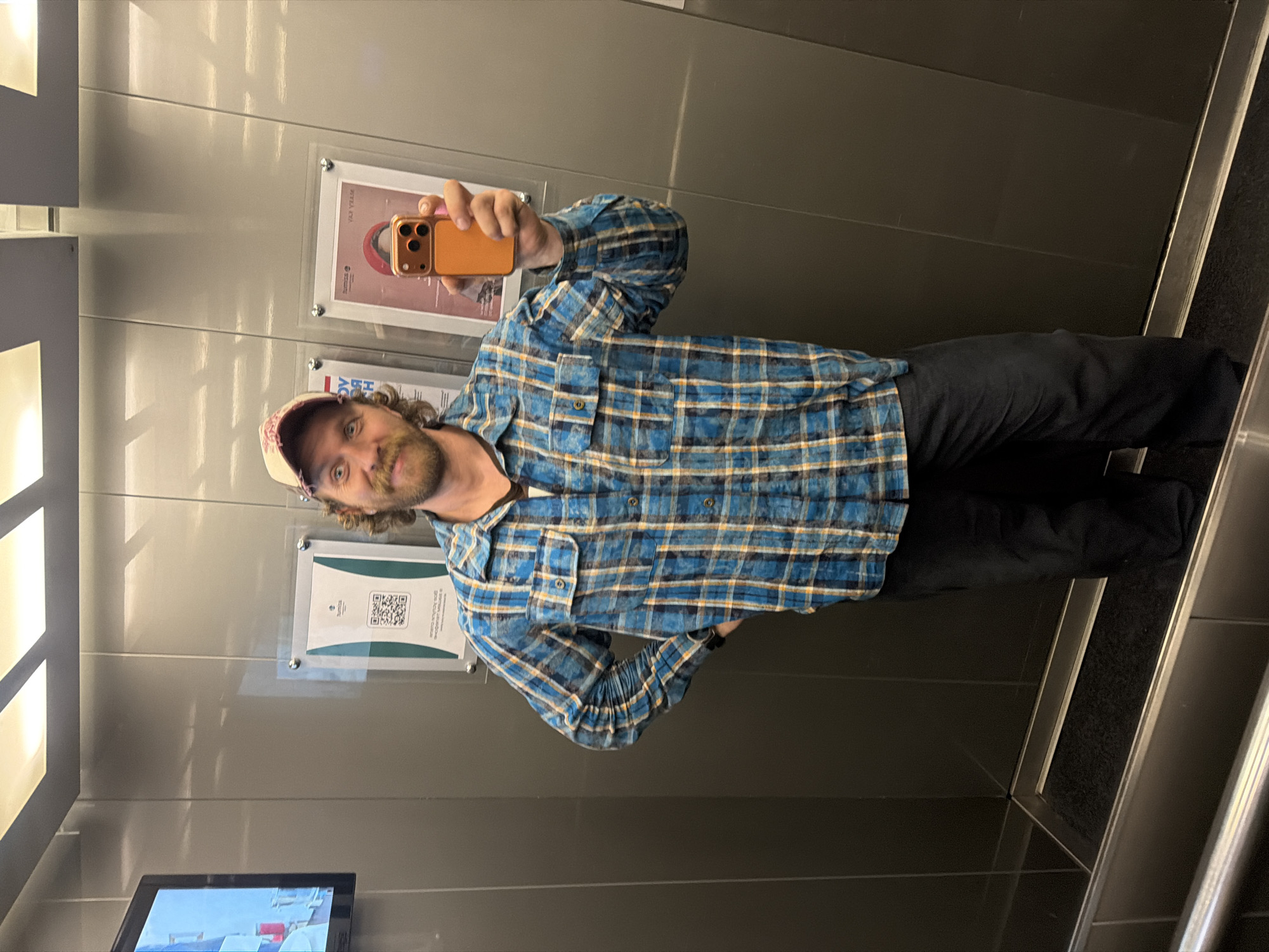 A man is taking a selfie in what appears to be an elevator. He is wearing a blue and yellow plaid shirt, dark pants, and a baseball cap. He is holding an orange phone in his right hand. Behind him are posters and a monitor. The elevator's interior is made of gray panels. The image was taken in Repino, Russia.