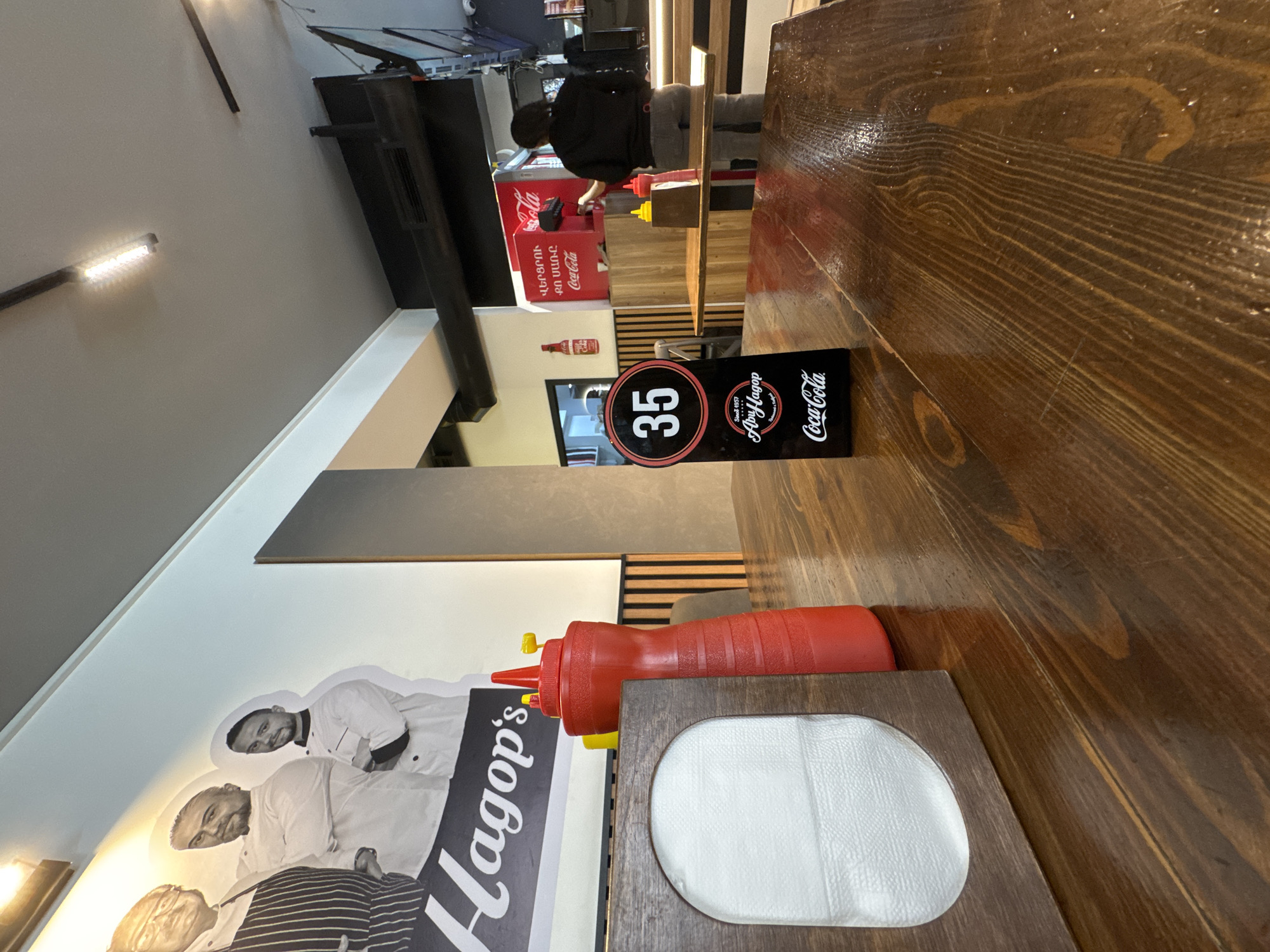 The image depicts the interior of Hagop's restaurant, a fast-food establishment in Yerevan, Armenia. A wooden table is prominent in the foreground, featuring a napkin holder, a squeeze bottle of ketchup, and a table marker with the number 35, along with Hagop's logo and the Coca-Cola brand. In the background, a person is standing near a Coca-Cola refrigerator unit. The wall features a graphic with images of men.