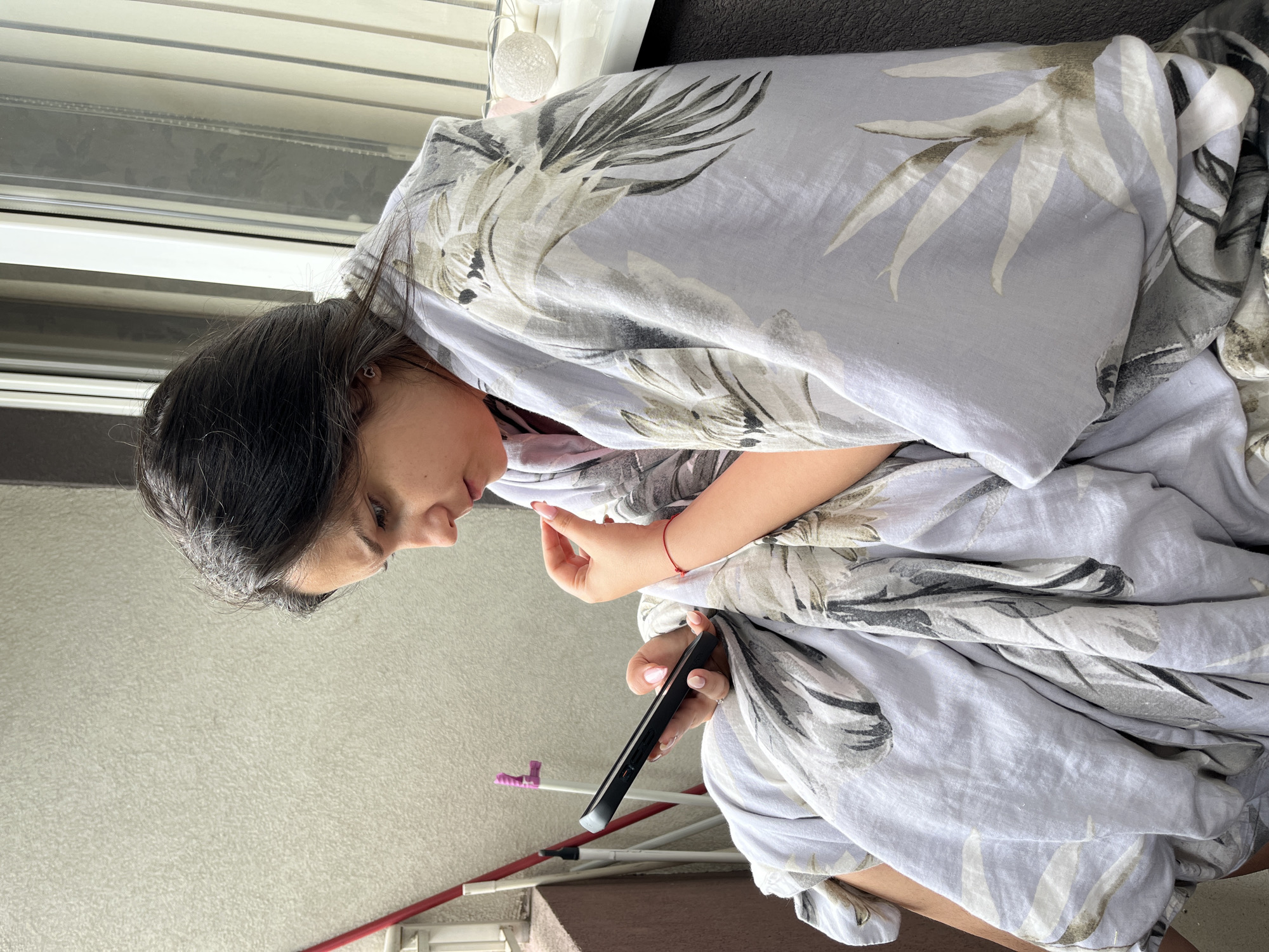 A young woman, with dark hair and fair skin, is seated on an outdoor balcony, wrapped in a light purple blanket with a palm leaf pattern. She is looking down at her black smartphone, which she holds in her left hand. Her right hand is raised, with her index finger touching her chin. A thin red bracelet is visible on her right wrist. The balcony has a textured, light-colored wall to the left and a dark grey surface to the right. A white-framed window with vertical blinds is visible behind her. A red pole, possibly part of a drying rack, stands to the left of the frame. The scene is likely located on a residential balcony in Sofia, Bulgaria.