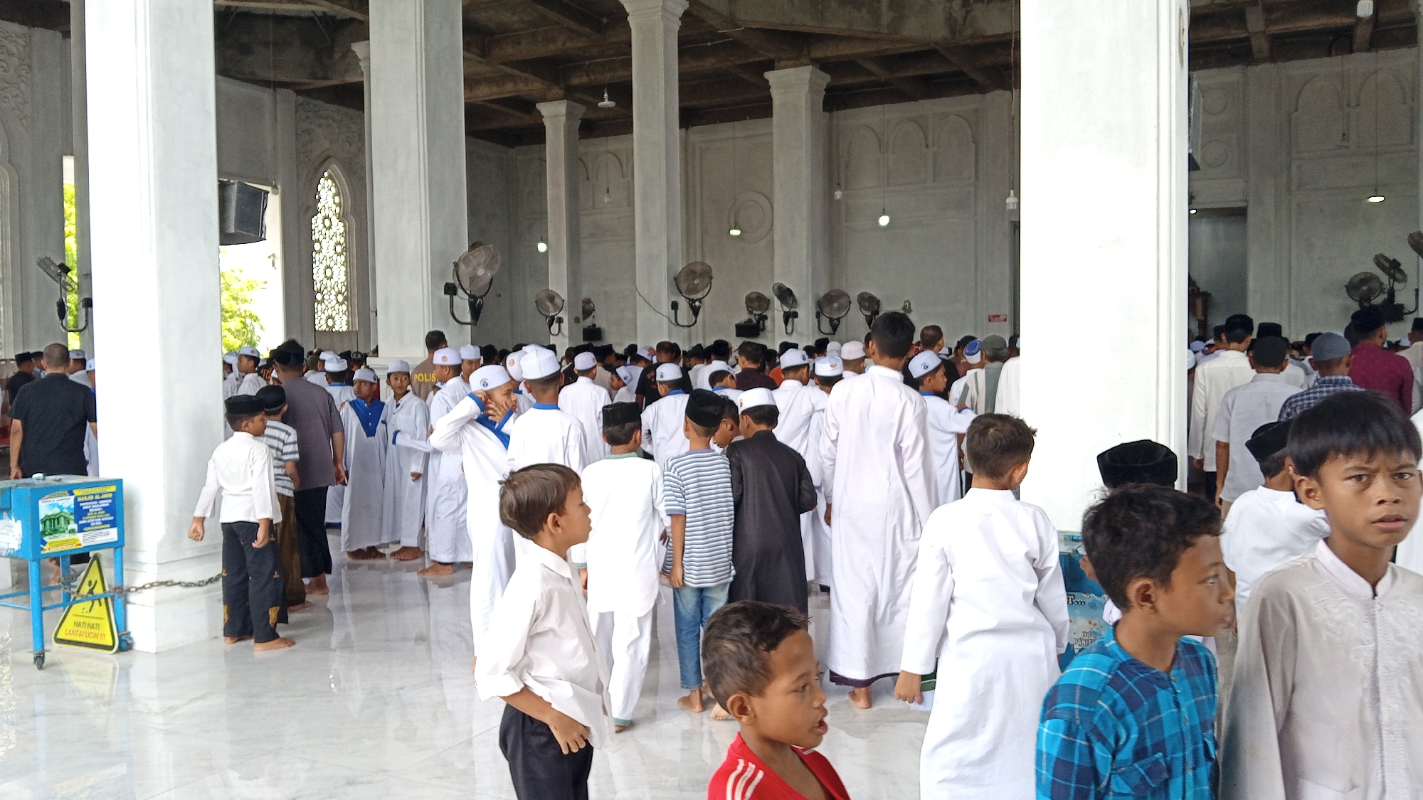 The image shows a group of people, mostly young boys dressed in white Islamic attire, standing inside a mosque. The mosque appears to have a large open space with tall pillars and fans mounted on the walls. The floor is tiled and reflective. In the foreground, several children are visible, some looking at the camera. In the background, more people, including adults, are gathered. There is a blue metal cart with some signs on it near the left side of the image. The overall scene suggests a religious gathering or event. The location is identified as the Masjid Agung Syeikh Abdurrauf in the city of Bireuen, Indonesia.