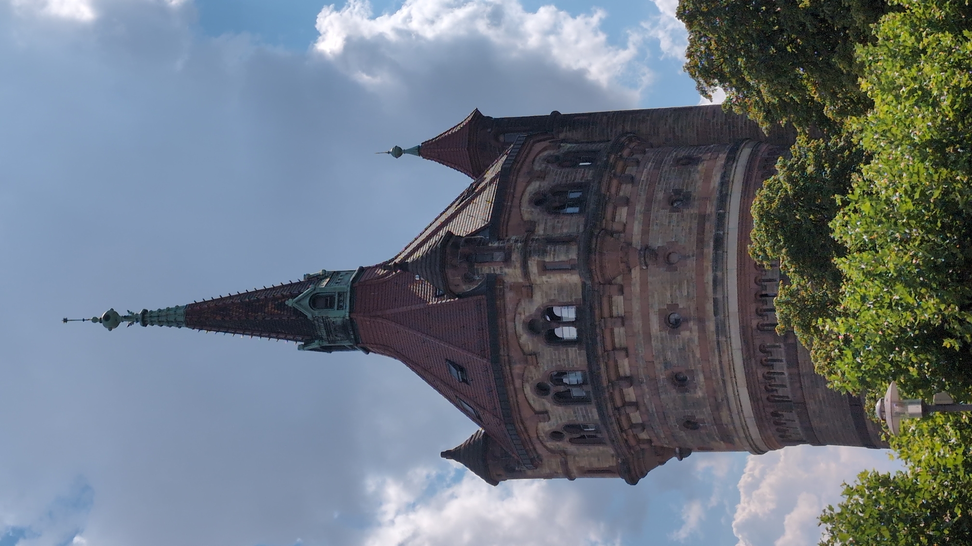 The image displays the historic Wasserturn (water tower) in Worms, Germany. The tower is constructed of brick with prominent red-tiled roofs on its various sections, including a tall, conical spire at its apex. It features arched windows and decorative architectural elements. The tower is partially obscured by lush green trees in the foreground, with a street lamp visible to the left. The sky above is a mix of blue with dramatic, fluffy white clouds. The overall impression is one of a historical landmark set against a backdrop of nature and sky.