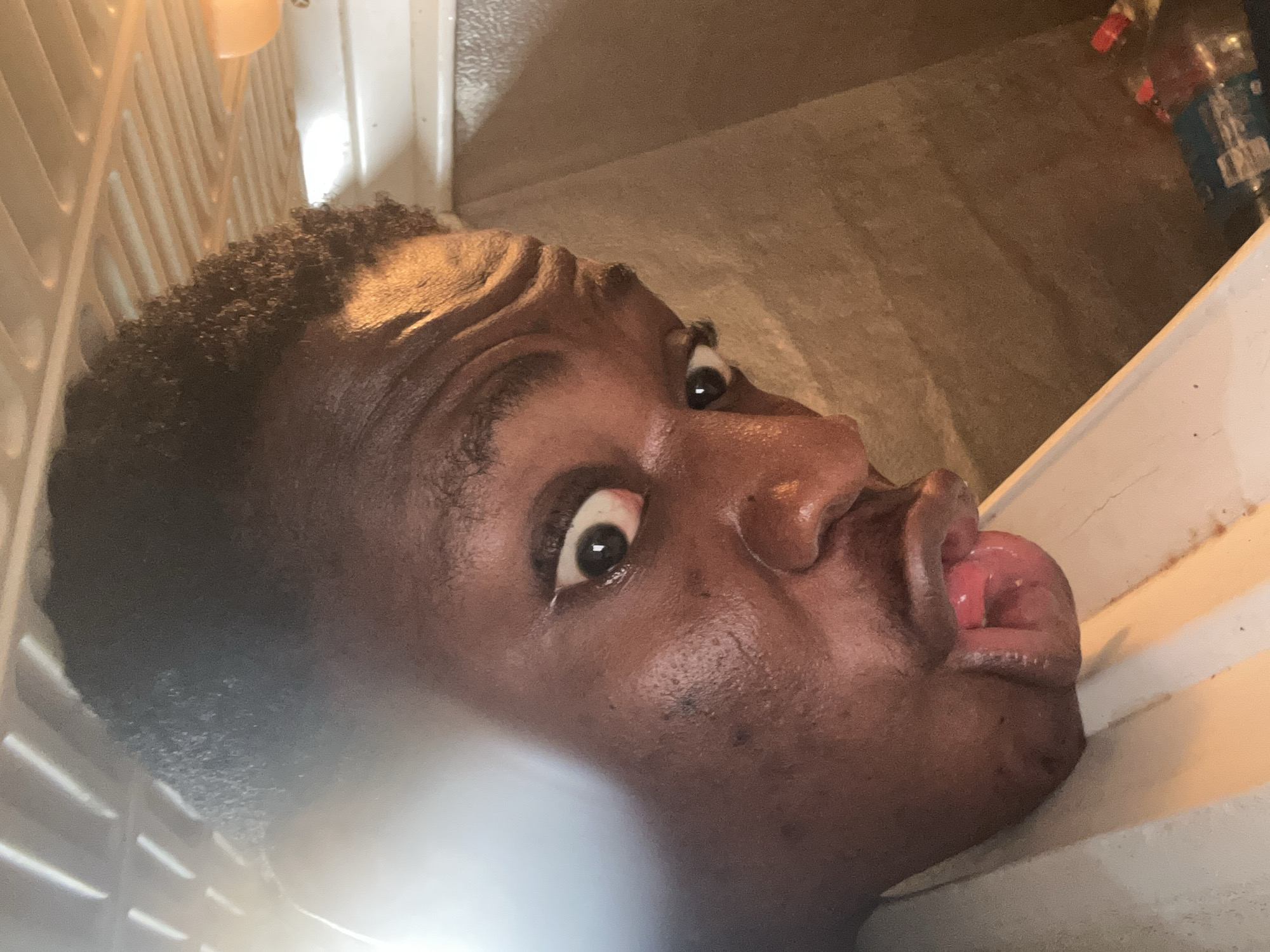 A close-up, tilted, low-angle shot depicts a dark-skinned person's face inside of a refrigerator. The individual's eyes are wide and appear startled. Their mouth is open with their tongue protruding. The refrigerator's white shelving is visible to the left of the face. A shelf with water bottles is visible behind the face, on the right. The location is not determinable from the given image.