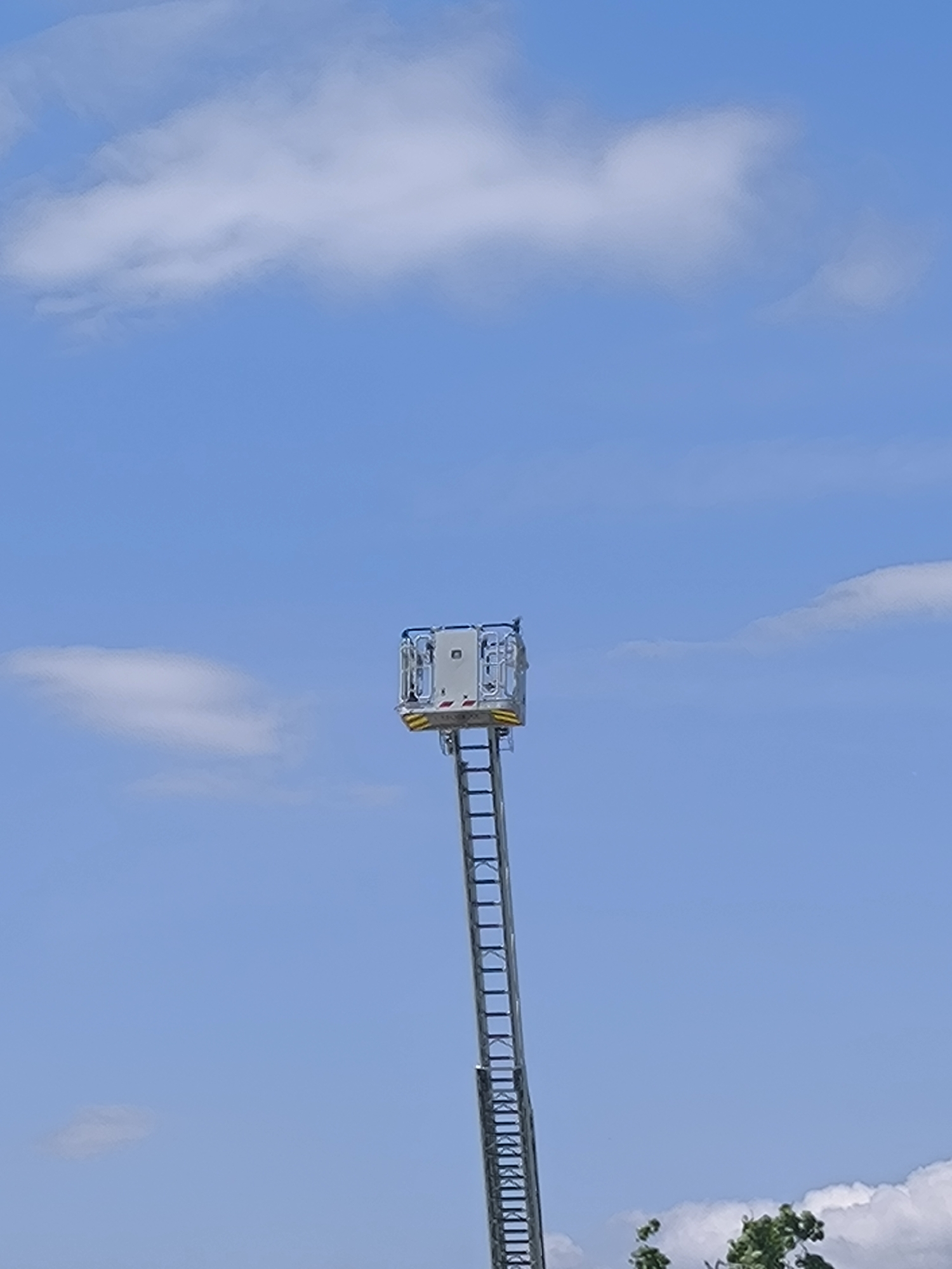 A low-angle shot depicts an extended fire truck ladder against a blue sky with scattered white clouds. The ladder extends upwards with a safety cage at its top. The scene is outdoors and details regarding the location (city, state) cannot be inferred from the image.