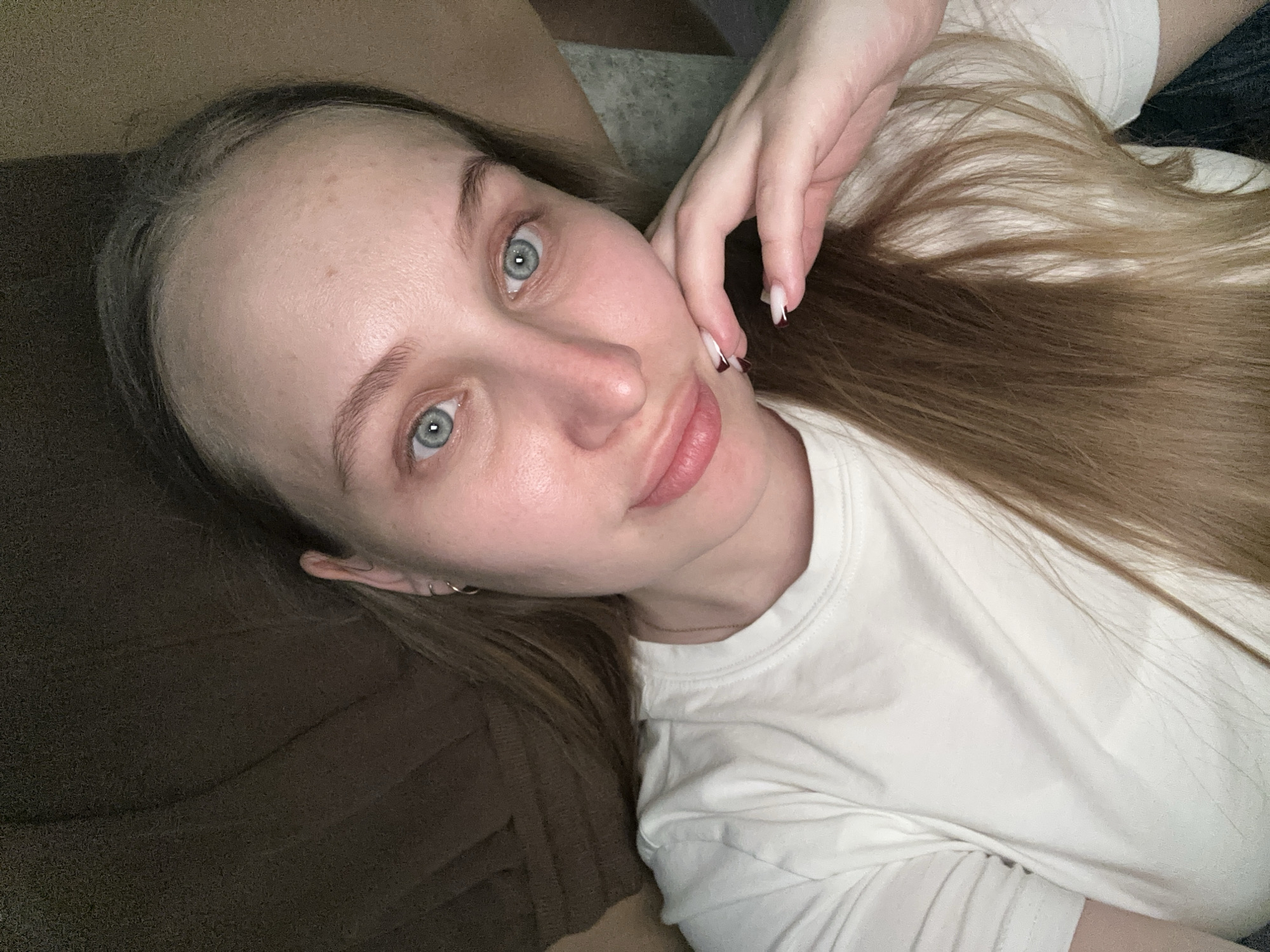 A young woman lies on what appears to be a brown sofa or cushion, wearing a white T-shirt. She's tilted her head towards the camera, her hand near her face. Her skin tone is fair, and she has light brown hair. The image suggests an indoor setting. There is no information in the image to suggest that this image was taken in Chaikovsky, Russia.