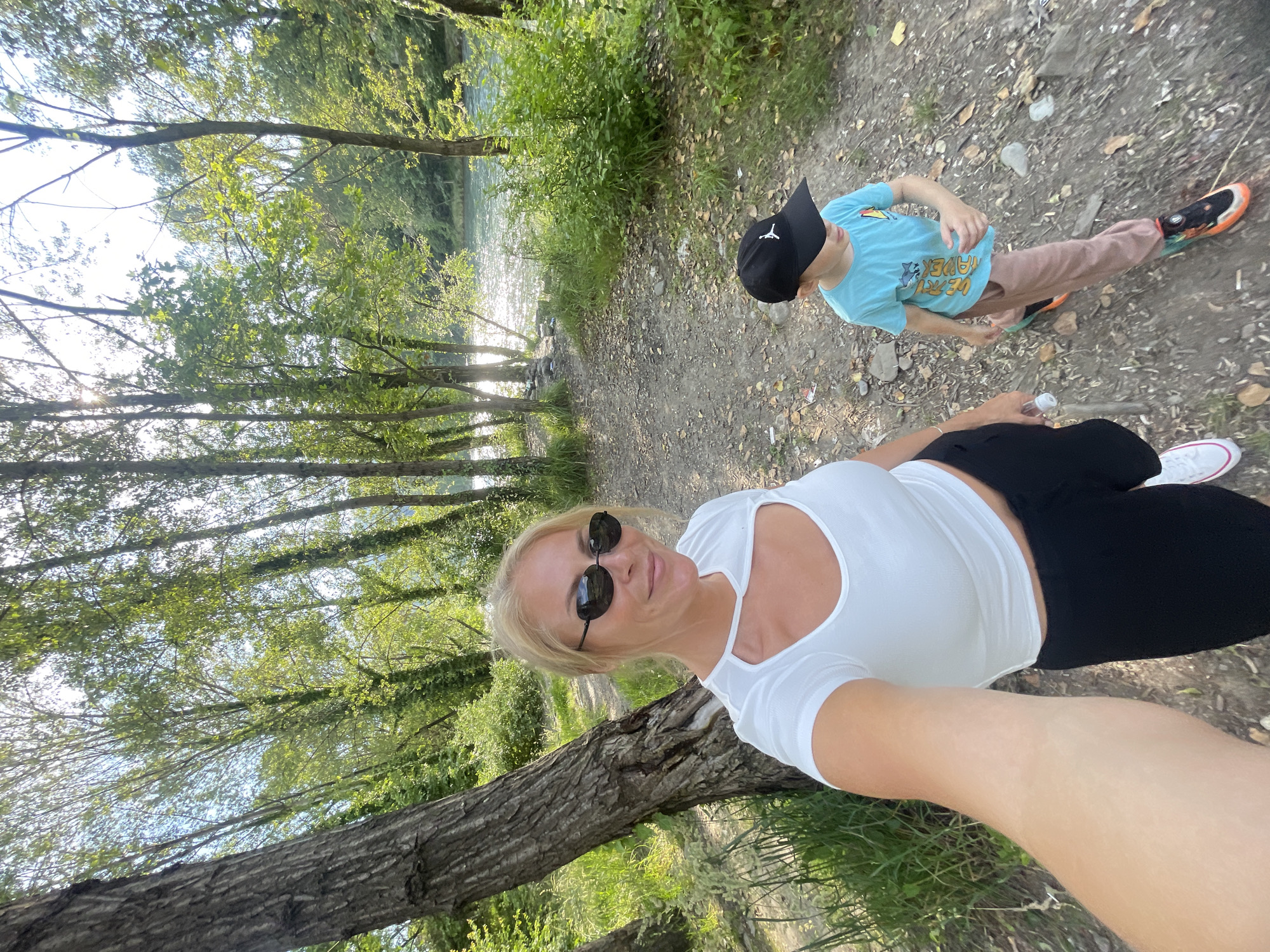 This selfie-style, outdoor, full-body shot shows two people on a dirt path near a body of water. The primary subject, a blonde woman with sunglasses, is in the foreground, wearing a white top and black pants. Her arm is extended, holding the camera. Behind her, a young boy with a black baseball cap and a light blue t-shirt is walking away from the camera. He's wearing tan pants and colorful sneakers. The path is lined with trees and lush green vegetation, and the water glints in the sunlight, suggesting it's daytime and likely fair weather. The overall mood is casual and active, hinting at a leisurely walk or hike. The word "MAVERICK" is visible on the boy's t-shirt. The location is contextually identified as Sochi, Russia.