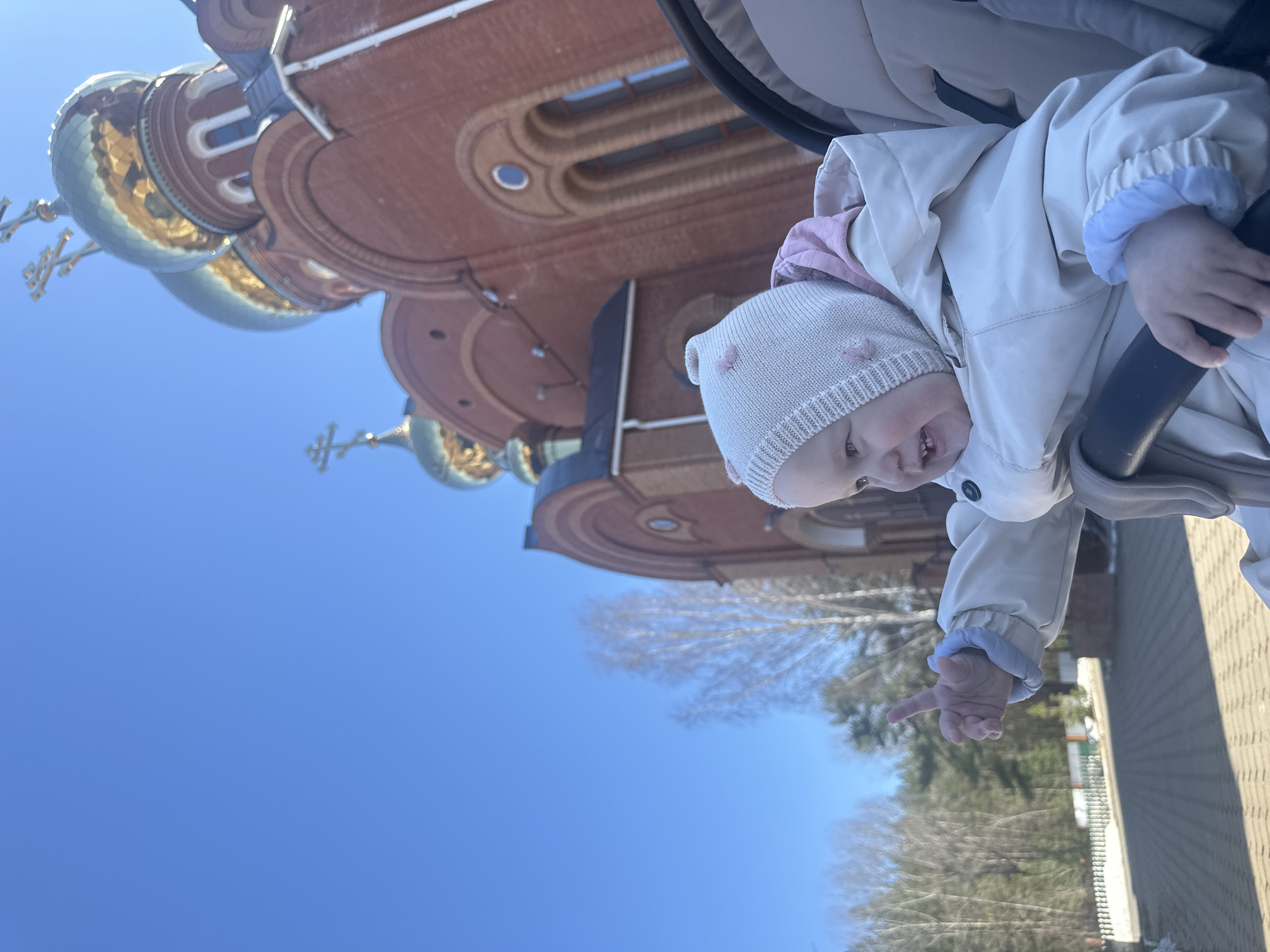 A baby in a white hat and beige jacket is in a stroller in front of the Alexander Nevsky Cathedral in Chaikovsky, Russia. The cathedral is red brick with golden domes topped with crosses. The sky is blue and clear. Trees are visible in the background. The baby is pointing with its right hand.
