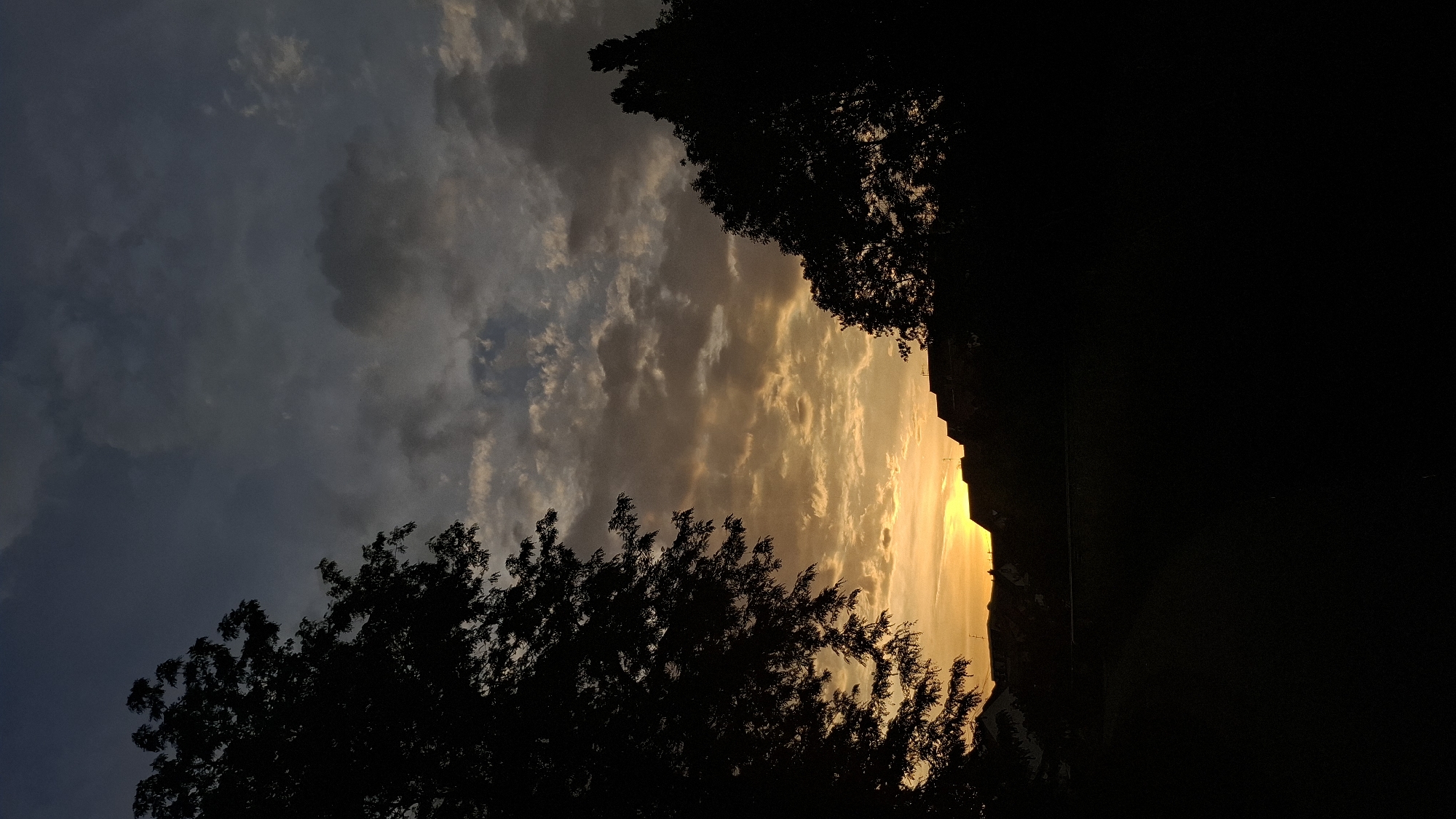 The image depicts a silhouette of a sunset scene. In the foreground, the silhouettes of trees and rooftops are visible against a vibrant sky. The sky is filled with clouds, illuminated by the warm hues of the setting sun, ranging from yellow to orange and darker shades of gray. The overall scene is dark, with the exception of the bright sky, suggesting either early morning or late evening. There is no visible indication of the city of Strasbourg, France, or any people in the image.