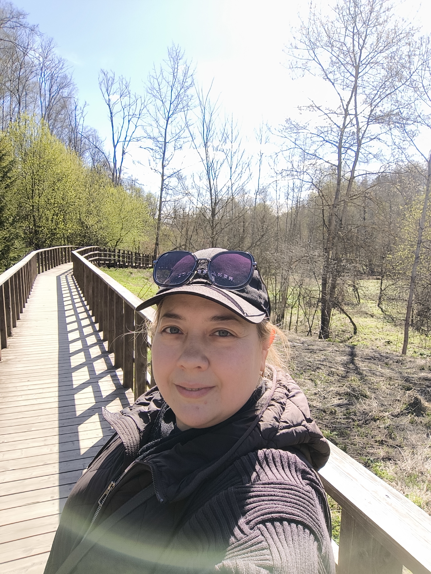 A woman captures a selfie on a bright, sunny day while traversing a winding wooden boardwalk through a natural park setting, likely in Moscow, Russia. She stands in the foreground, facing the camera with a gentle smile. She has light-colored eyes and is wearing a dark baseball cap, with large, dark, possibly purple-tinted sunglasses resting on top. A reversed reflection of text, appearing as "SVEZ", is visible on the sunglasses' lens. Her attire consists of a dark, possibly quilted, jacket worn over a dark sweater featuring a distinctive ribbed pattern on the sleeves and shoulder.
The wooden boardwalk, equipped with railings, extends and curves into the background on the left side of the frame. Strong, diagonal shadows from the railings are cast across the boardwalk's planks, indicating bright sunlight and suggesting a mid-morning or early afternoon time. To the left of the boardwalk, a hillside is densely covered with trees and shrubs exhibiting fresh, light green spring foliage. To the right, a flatter area shows patchy green and dry grass, interspersed with numerous deciduous trees that are either bare-branched or just beginning to bud. The background is dominated by a clear, bright blue sky with faint wisps of clouds, completing a scene of early spring in a tranquil park.