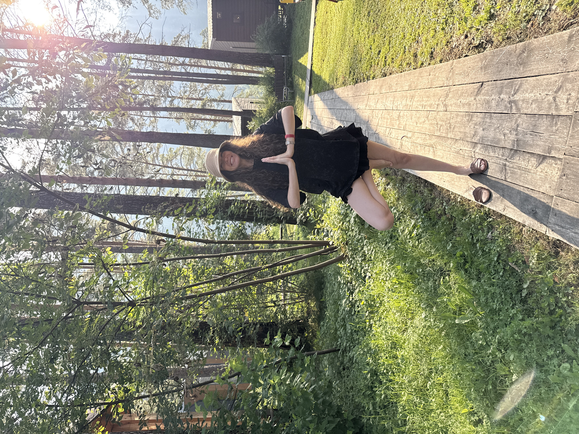 In Barangol, Russia, a woman stands in a tree pose on a wooden walkway amidst a forest. She wears a black t-shirt, black shorts, a beige hat, and brown sandals. Her hands are pressed together in a prayer position at her chest. The sun filters through the trees, casting shadows on the ground. To the left of the walkway is lush green grass and trees, while to the right are more trees and some small wooden structures. The background suggests a natural, possibly rural or resort setting.