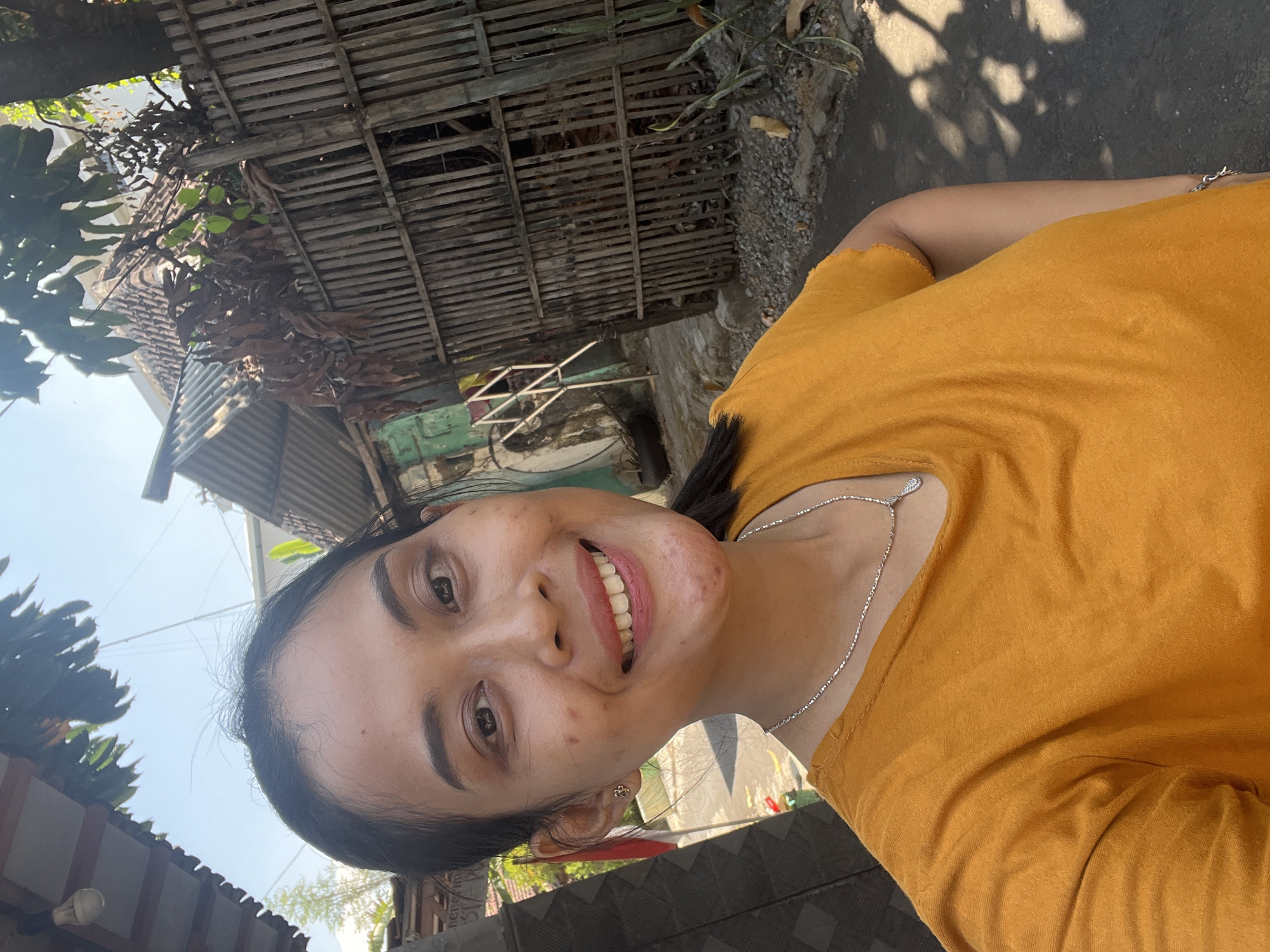 A young Indonesian woman of East Asian descent, with dark hair pulled back and a visible skin condition on her face, smiles into the camera. She is wearing an orange t-shirt and a silver necklace with a pendant. The background suggests a residential area, possibly in Kecamatan Pati, Indonesia, with a bamboo fence, corrugated metal roofing, and some greenery. The scene is outdoors, under a clear sky, with dappled sunlight creating shadows on the ground.