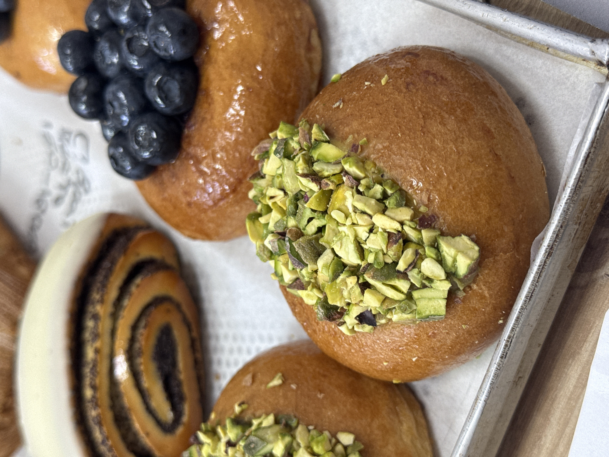 The image displays a close-up, high-angle view of various pastries on a baking tray lined with white paper. Several round, golden-brown buns or donuts are visible.
In the upper left, one glossy bun is generously topped with a cluster of fresh, dark blue blueberries. To its right and in the foreground, another bun features a substantial topping of chopped green pistachios. A third, partially visible bun in the bottom left also appears to be topped with pistachios. On the far left, a distinct spiral-shaped pastry, possibly a poppy seed roll, is visible, glazed with white icing on one side. The pastries have a shiny, possibly glazed finish, suggesting sweetness.
The scene takes place indoors, likely in a bakery or kitchen, given the presence of a metal baking tray. No specific activity is captured, but the arrangement suggests a display or preparation of freshly made sweet treats.
Notable details include the varied toppings on the pastries, providing contrast in color and texture. The lighting is bright and even, highlighting the golden hues of the buns and the vibrant colors of the toppings. No people are visible, and therefore no emotions can be discerned. There is no visible indication of the time of day or weather. While the location context is provided as Belgrade, Serbia, there are no visual cues within the image itself to confirm this. Blurry, illegible text, possibly a logo or brand name, is faintly visible on the paper liner near the blueberry-topped pastry.