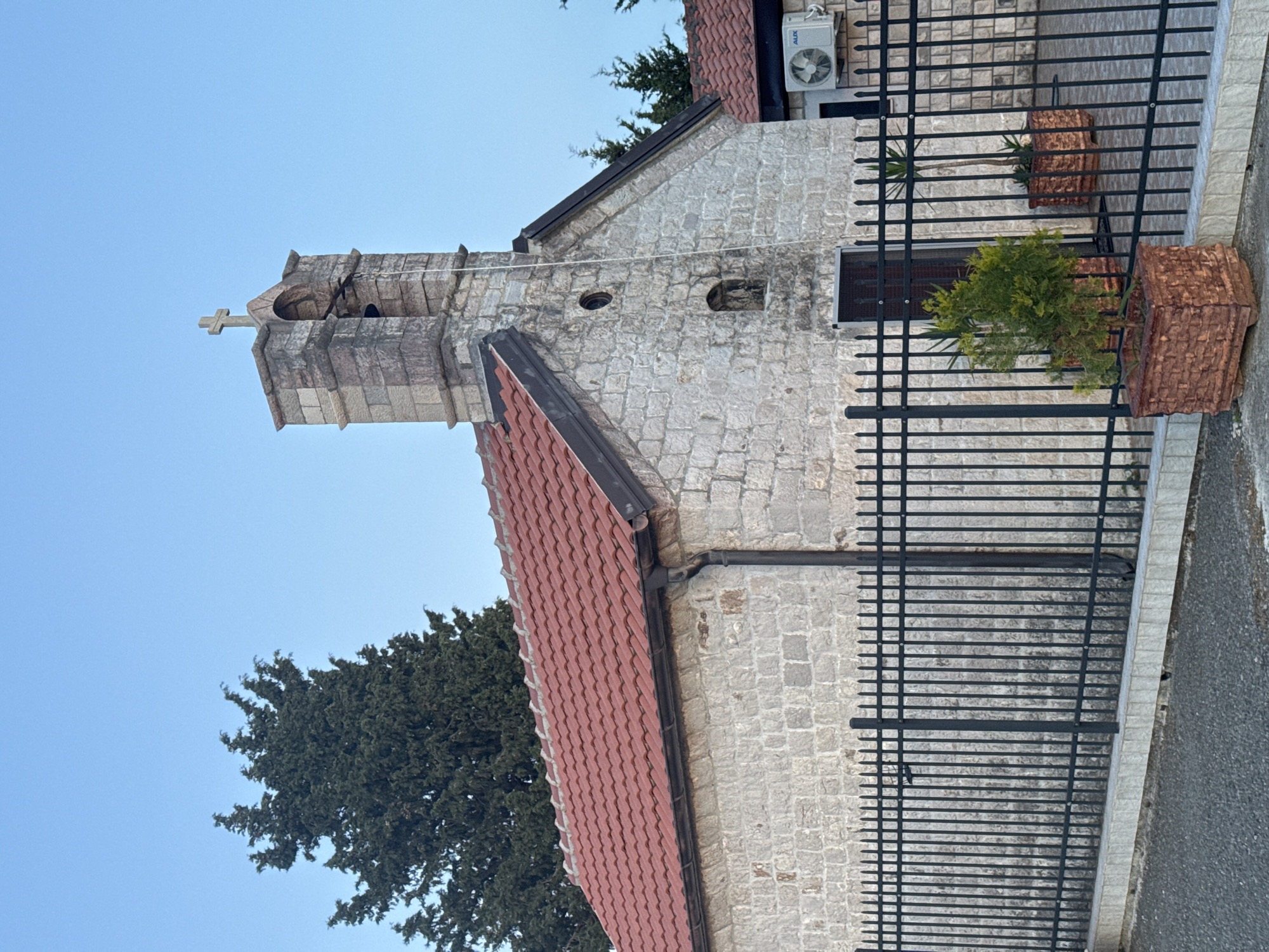 The image depicts a stone church in Golubovići, Montenegro. The church features a bell tower with a cross atop. It has a red-tiled roof and is surrounded by a black metal fence. Potted plants are visible behind the fence. An air conditioning unit is mounted on the building's exterior. A large tree is visible next to the church. The sky is clear and blue.