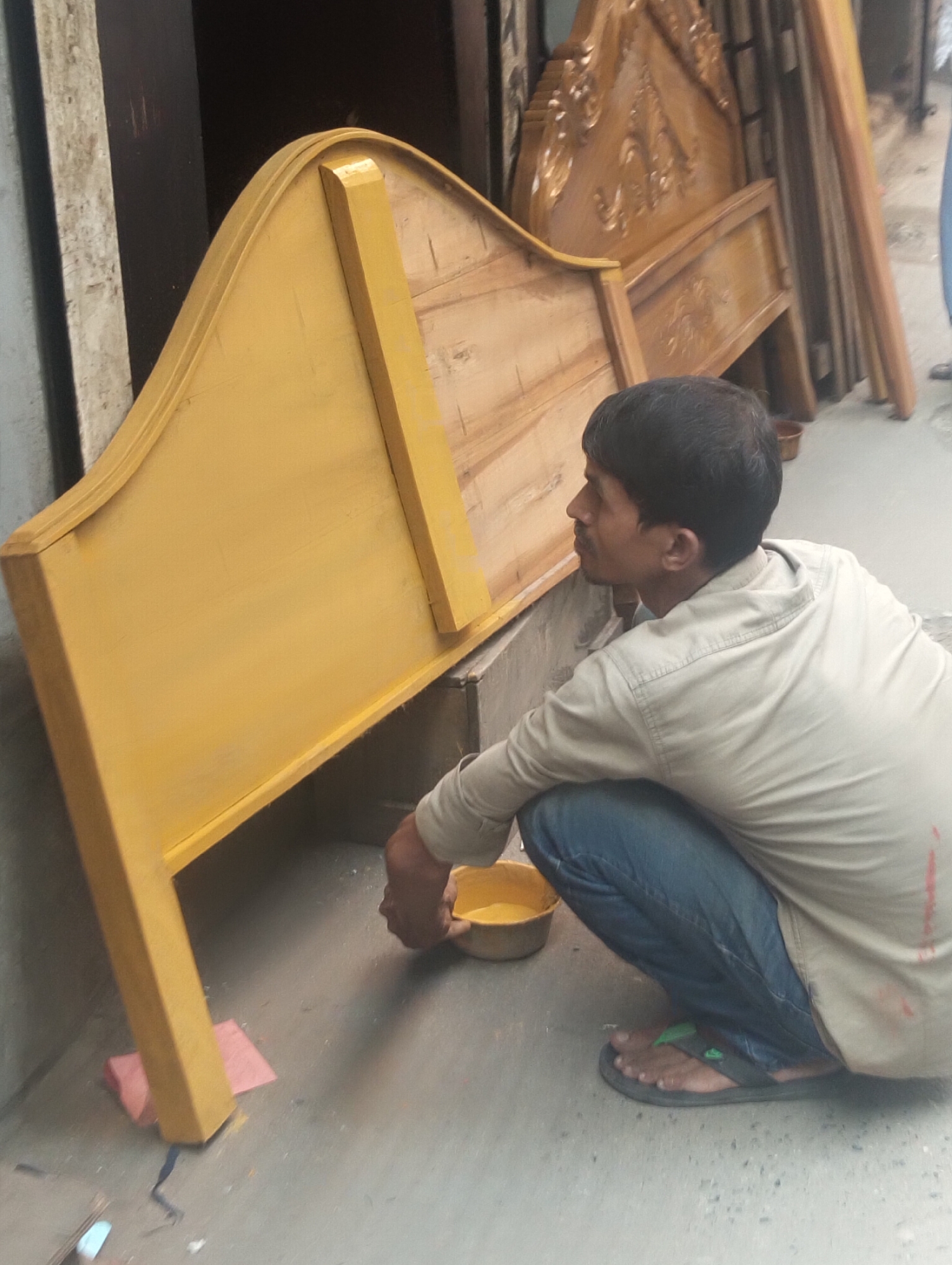 In Dhaka, Bangladesh, a man wearing a light-colored shirt, blue jeans, and sandals squats to paint a wooden headboard yellow. The headboard stands upright on a concrete surface, supported by a small piece of pink material. Another headboard, unpainted and more ornately carved, leans against a wall nearby. The man holds a small bowl of yellow paint. The environment appears to be a street or sidewalk.