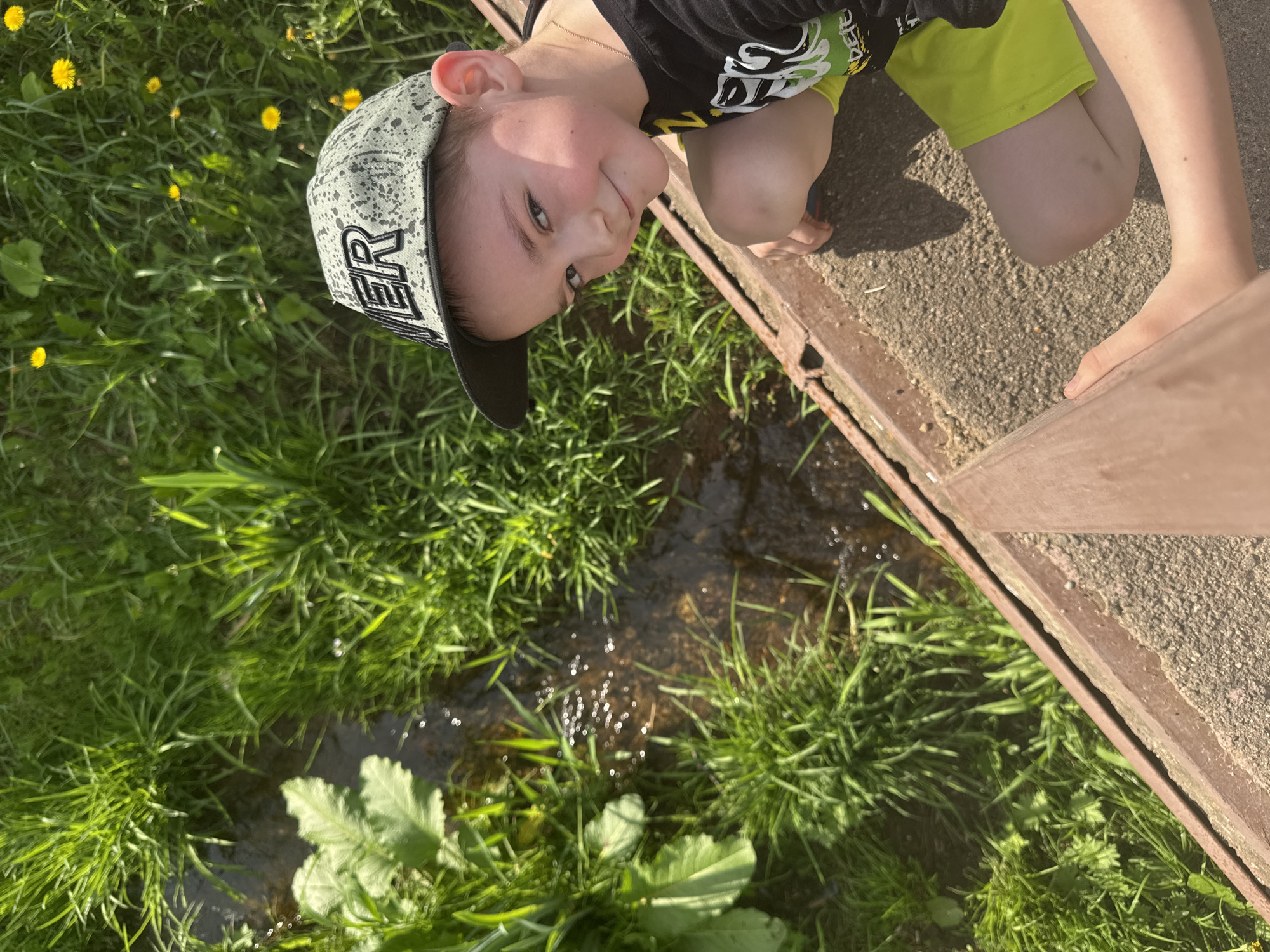 A young boy wearing a gray cap, black t-shirt, and yellow shorts is looking into a ditch in Shevlyakovo, Russia. He is crouched near the edge of a concrete and metal structure with green grass and dandelions on the other side. Water is visible in the ditch.