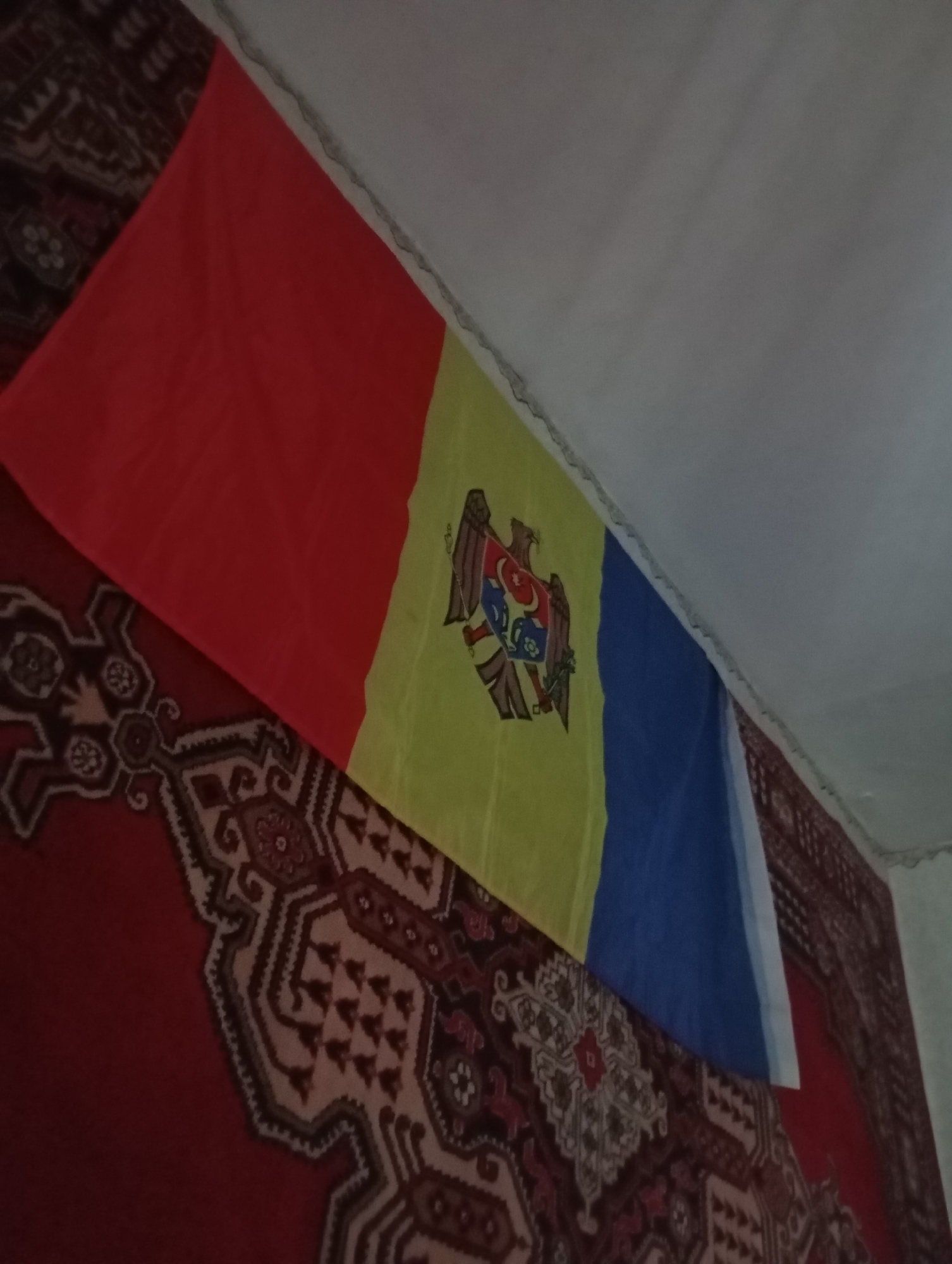 A flag is displayed on a wall. The flag has three vertical stripes of red, yellow, and blue, with the Moldovan coat of arms on the yellow stripe. The flag is hanging on a wall, and a portion of a carpet with a red and dark patterned design is visible at the bottom of the image. The scene appears to be indoors, possibly in a room. No people or actions are depicted. The lighting is dim. The specific location in Soroca, Moldova, cannot be determined from the image alone.