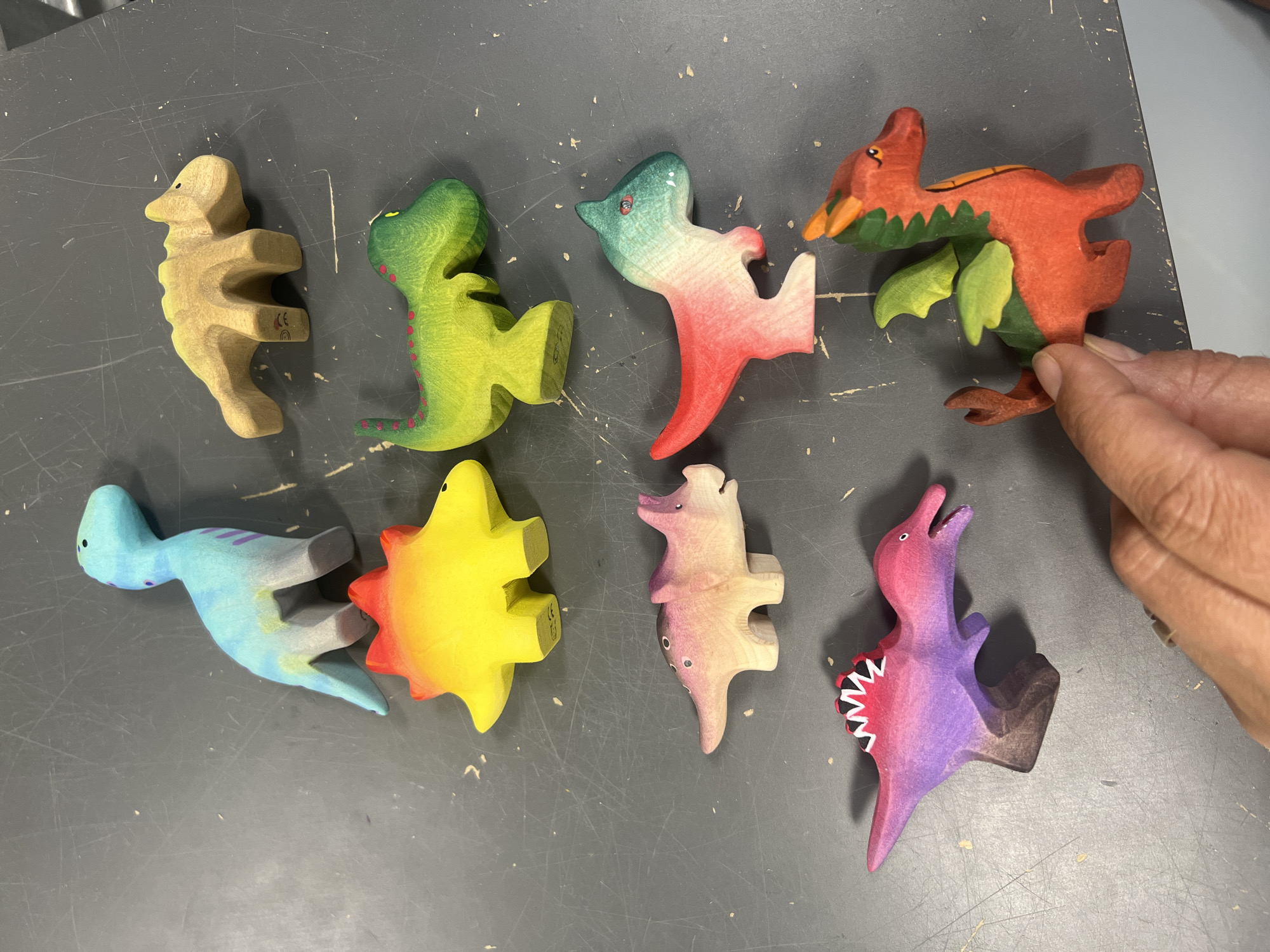 The image displays a collection of eight wooden toy dinosaurs arranged on a dark gray, textured surface. The toys are brightly colored and have a smooth, finished appearance. From left to right, top row: a light brown, abstractly shaped dinosaur; a vibrant green dinosaur with red spots; a dinosaur with a gradient of blue, pink, and red; and a red and green winged creature that appears to be a pterodactyl. Bottom row: a light blue dinosaur with purple stripes; a yellow and orange star-shaped object, possibly a sun or a stylized creature; a pink and purple dinosaur with dots; and another pink and purple dinosaur with a crest of white and black teeth. A person's hand is visible on the right side of the frame, gently holding the pterodactyl. The lighting suggests an indoor setting, possibly a craft or play area. There are no explicit time of day or weather cues. The overall scene is focused on showcasing the colorful and handcrafted nature of the wooden toys. Some of the toys have small markings on their undersides, including a "CE" symbol and a rainbow icon on the brown dinosaur and the star object, respectively.