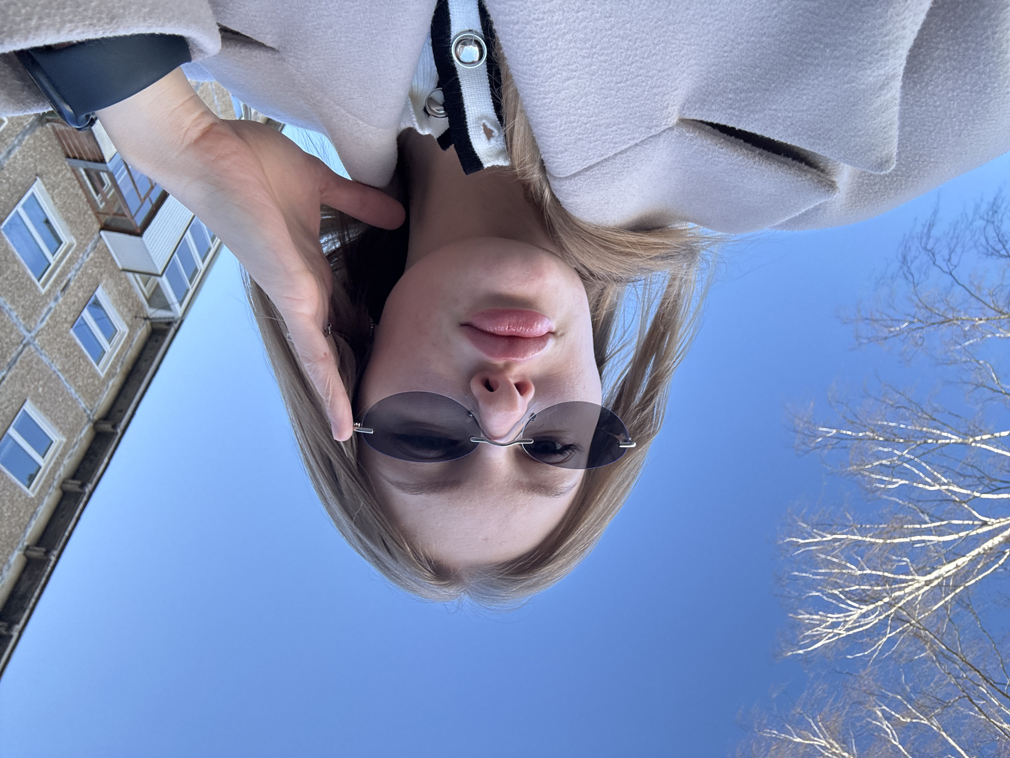 A selfie taken from a low angle shows a blonde woman wearing sunglasses and a light-colored coat with a black and white trim. The background consists of a clear blue sky, bare tree branches, and a multi-story building with visible windows. The picture was taken outside in daylight. The location might be Perm, Russia, as the city was explicitly mentioned.