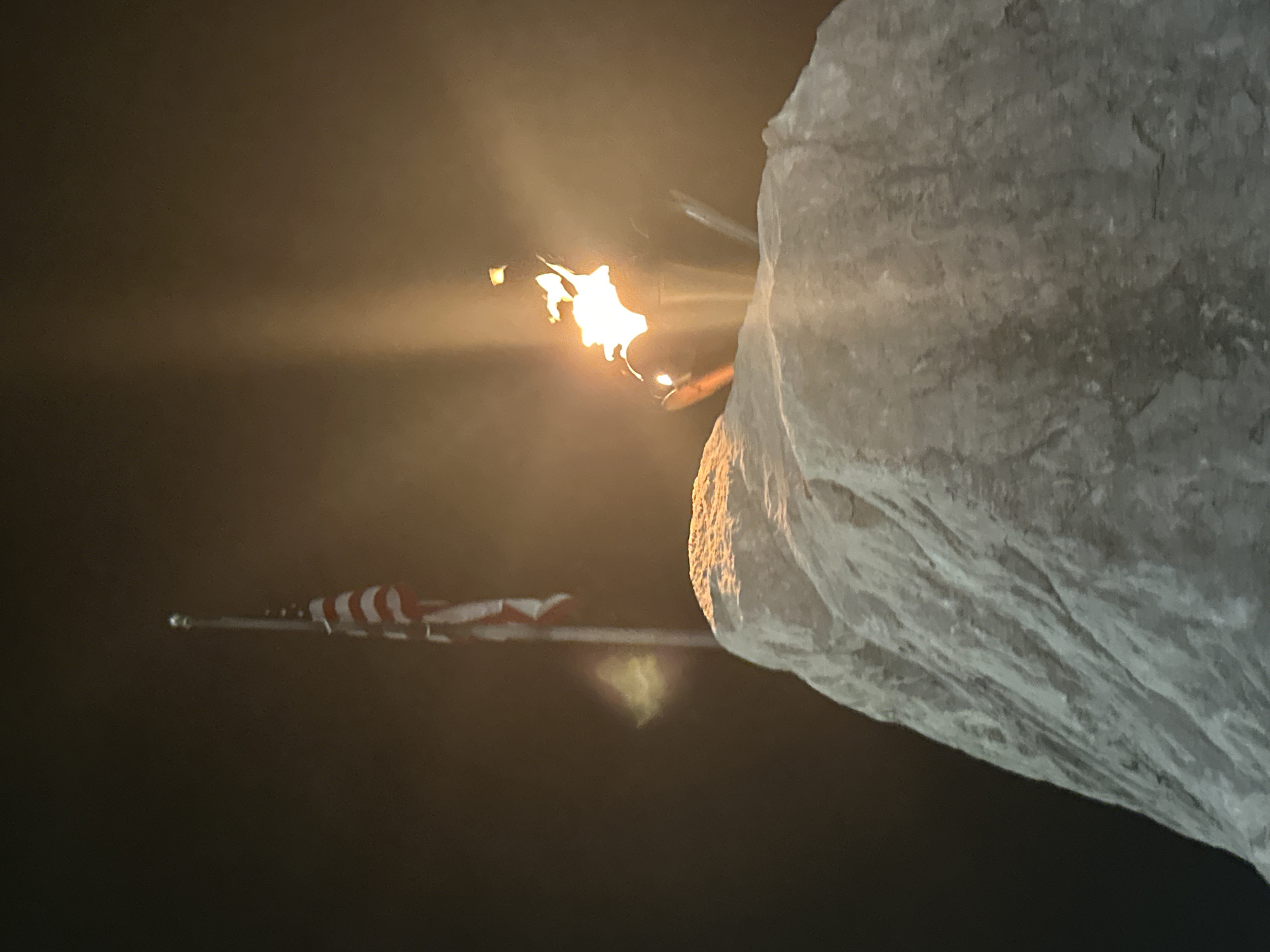 A close-up, low-angle shot captures a torch burning brightly on top of a large, rough-textured rock. The flame emits a strong glow, casting light and creating lens flare effects in the dark, hazy atmosphere. Behind the rock and to the left, an American flag flies on a tall flagpole. The flag is partially visible, with its red and white stripes evident. The overall scene is dark and atmospheric, suggesting it might be nighttime or a very overcast day, possibly a memorial or a scenic overlook in Huntsville, Alabama, given the presence of the American flag and a prominent rock formation.