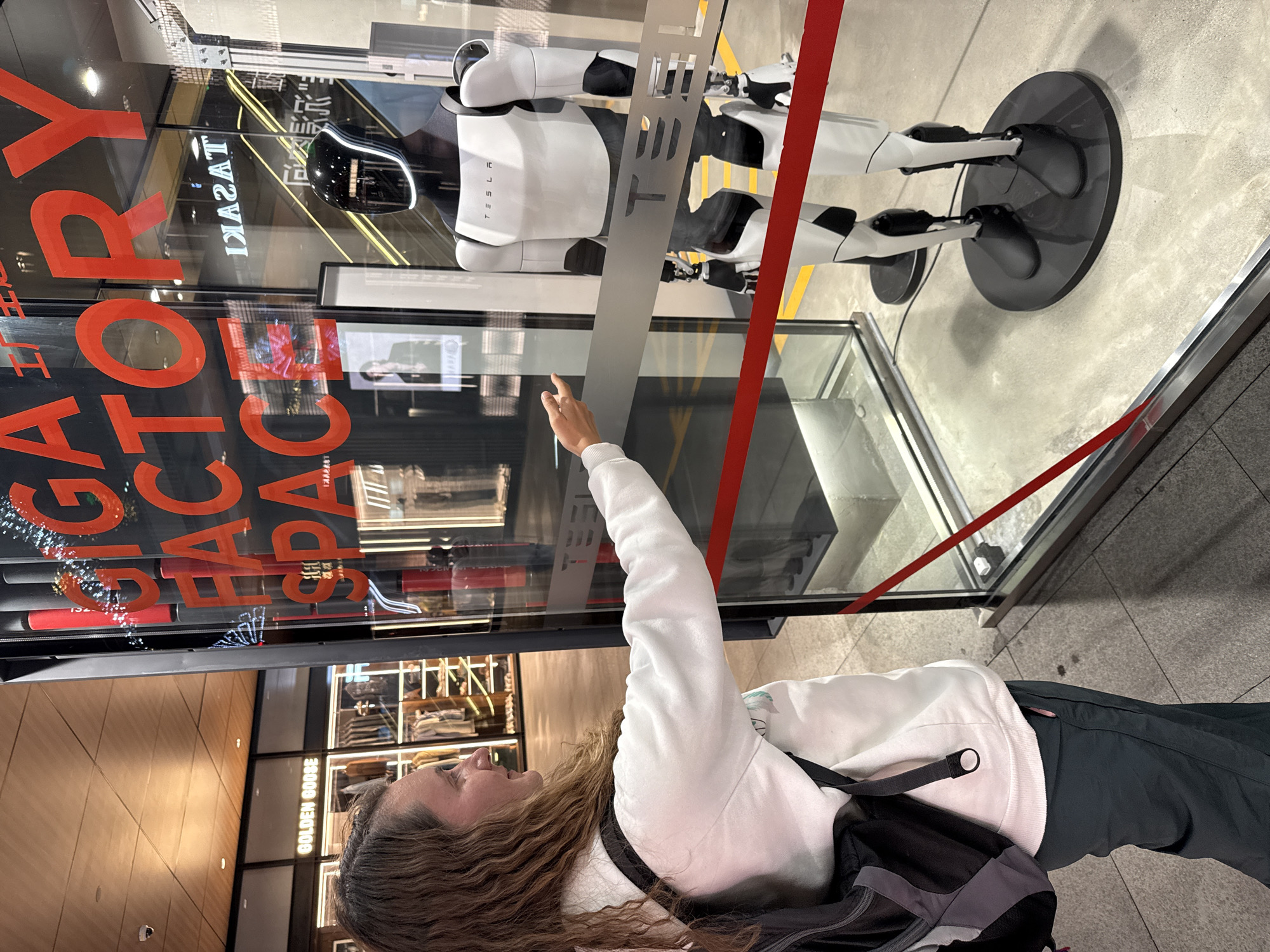 A person with long curly hair, wearing a white sweatshirt and black pants with a black backpack, points their right index finger towards a Tesla Optimus robot displayed in a glass enclosure. The robot is predominantly white with black accents, standing on a circular black base. The display is located in a storefront window with red lettering spelling "GIGA FACTORY SPACE" visible on the glass. The reflection in the window reveals the interior of a clothing store named "GOLDEN GOOSE". The scene is in Jinjiang District, Chengdu, China, indicated by the shop name "TASAKI" written in Chinese characters on the top left corner of the glass enclosure. The ground outside the enclosure is paved with tiles. A red tape stretches across the display.