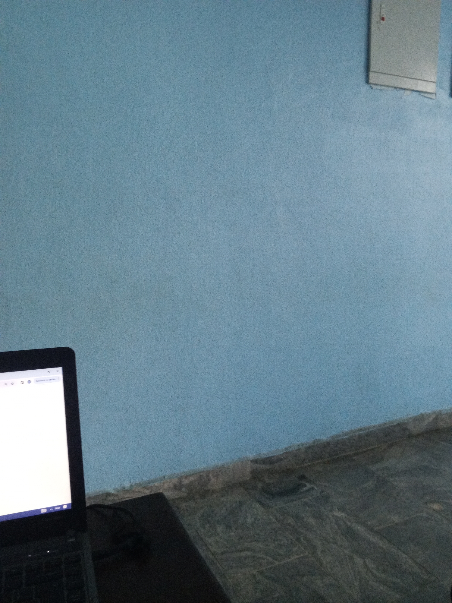 In Bori, Nigeria, a laptop sits on a dark surface in front of a pale blue wall. The laptop screen is illuminated, displaying what appears to be a web browser with a white page. To the upper right of the wall, a light-colored electrical panel is mounted. The floor below the wall is tiled with a dark, marbled pattern. The scene is illuminated by ambient light, creating a slightly dim atmosphere.
