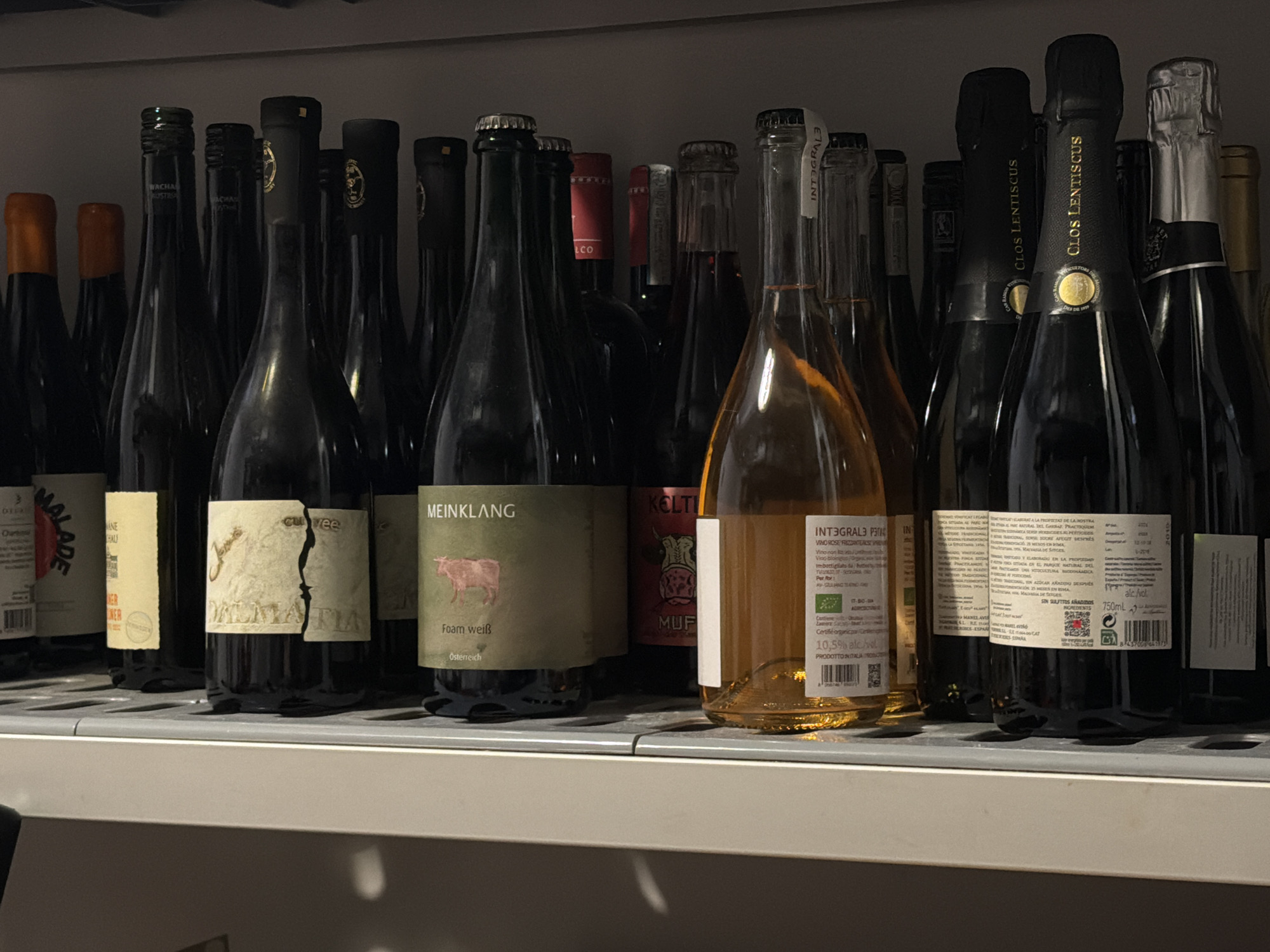 The image shows a shelf stocked with various bottles of wine. The bottles are arranged closely together, with labels partially visible. Some labels indicate brands such as "MEINKLANG," "CLOS LENTISCUS," "INTEGRALE," "ALADE," "KELTI," and "Cuvee DALMATIA." Details on the labels include information about the wine type, origin, and alcohol content. The bottles are of varying shapes and sizes. The shelf appears to be made of a light-colored material. The overall lighting is somewhat dim, with shadows obscuring some of the details. There is no information in the image about the location in Belgrade, Serbia. There are no people in the image.