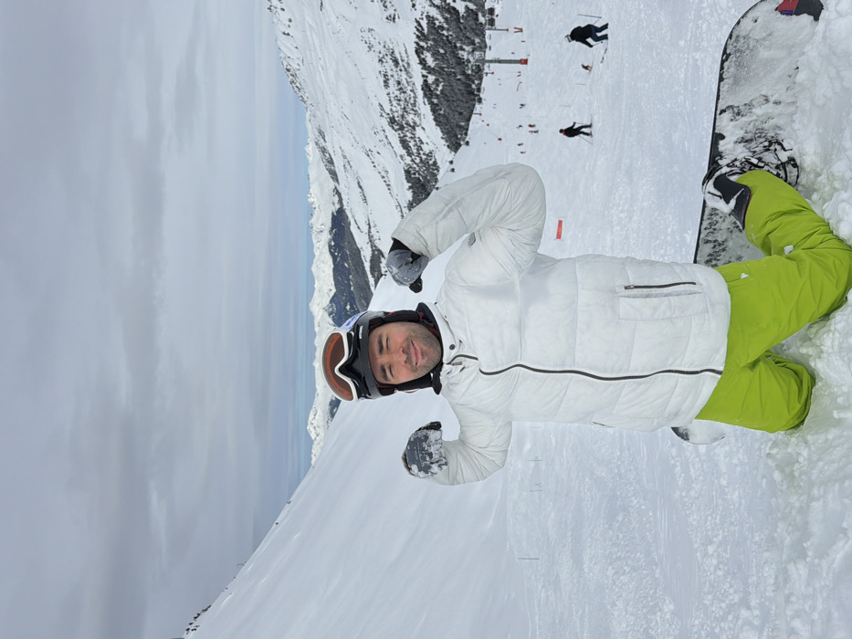 A male individual is depicted kneeling on a snowboard in a snow-covered mountain environment in Les Allues, France. The subject, facing the camera, wears a black helmet with orange-tinted goggles positioned on top, a white quilted puffer jacket with a black central zipper, neon green ski pants, and grey-white gloves. He is flexing both arms at the biceps, forming fists, and smiling. The snowboard is partially visible beneath him. The background features extensive snow-covered mountain terrain. To the left, a steep, smooth snowfield ascends. To the right, a ski slope with several skiers and snowboarders, appearing as dark figures, and multiple red ski lift poles are visible. Distant snow-capped mountain ranges stretch across the horizon under an overcast sky.