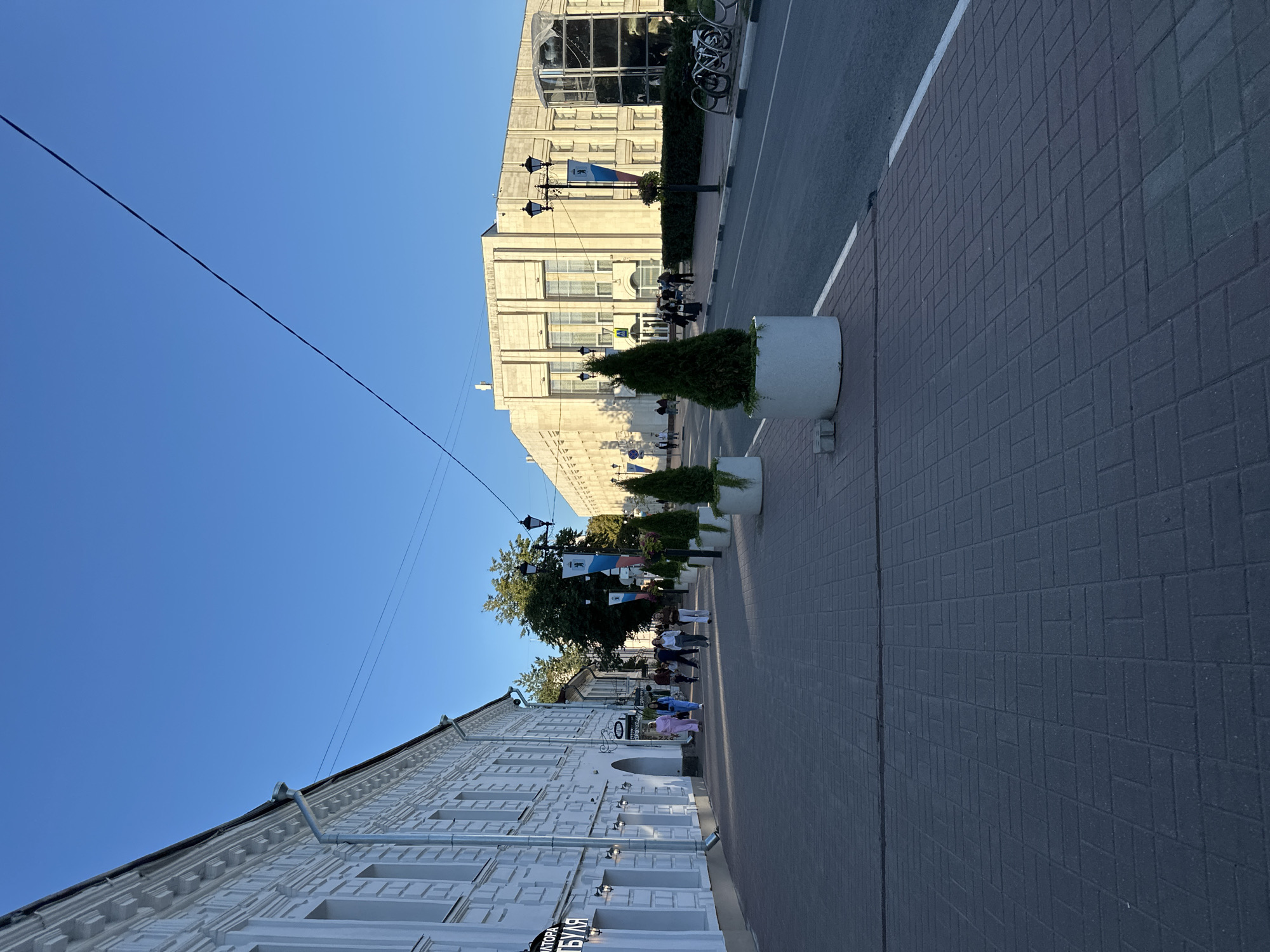 The image depicts a street scene in Yazvitsevo, Russia. On the left, a white building with ornate architectural details lines the sidewalk. Several people are walking on the sidewalk in front of the building. Across the street, a large, light-colored building with a classical facade stands under a clear blue sky. Planters with conical evergreen trees are placed along the sidewalk in front of this building. A road runs alongside the sidewalk, and there are bicycles parked near a modern glass-enclosed structure. Power lines are visible stretching across the sky. The overall environment suggests a daytime scene in a city.