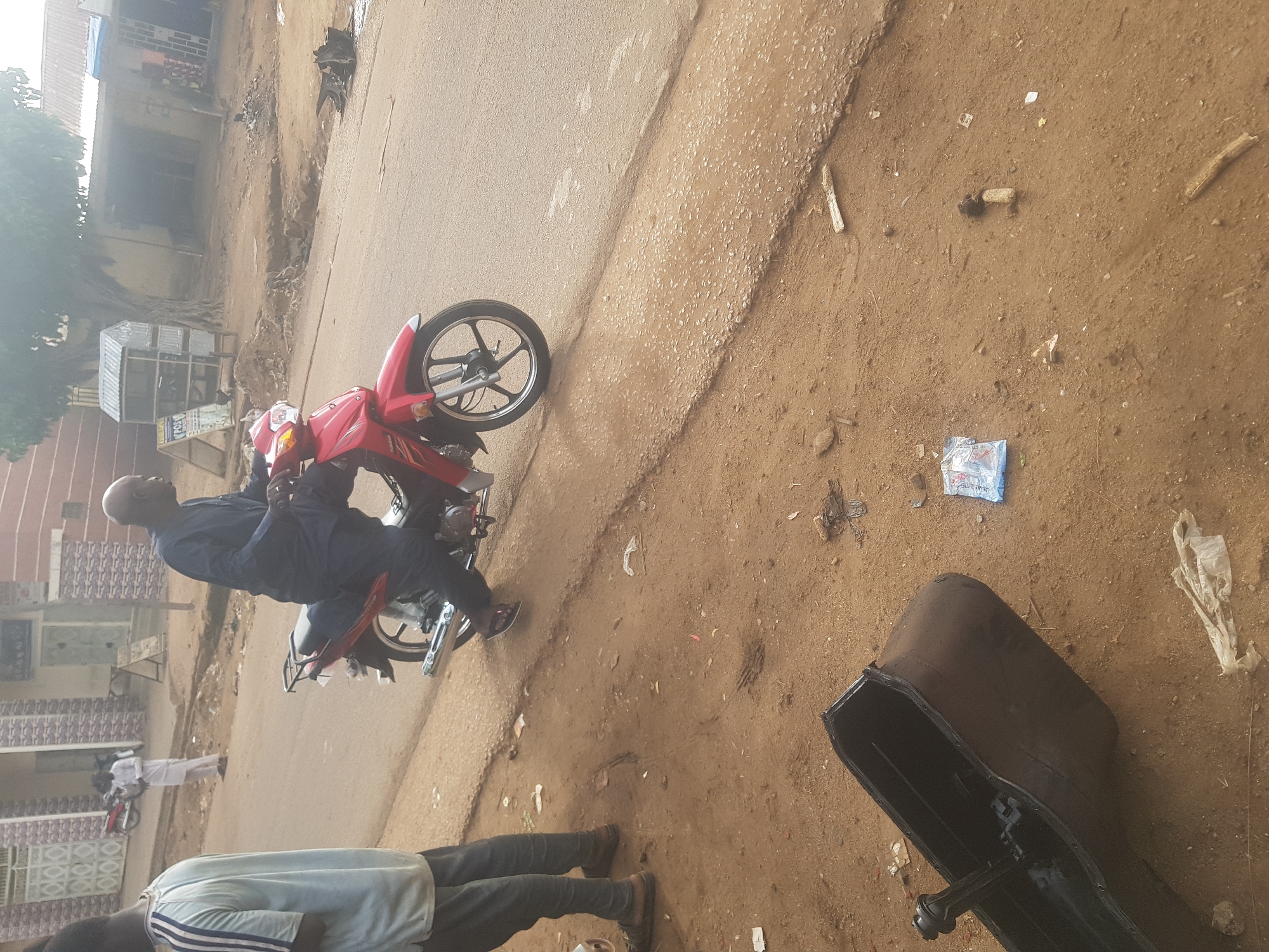 The image captures an outdoor street scene in Tukur Tukur, Nigeria, under bright daylight. The main subjects are three men, a red motorcycle, and a large black pan.
In the midground, a bald man wearing a dark blue long-sleeved shirt and trousers is seated on a red Honda motorcycle, which is stationary on a paved road. His feet are on the ground, suggesting he is either paused or about to move. The motorcycle faces towards the left side of the frame (if upright).
In the foreground, partially visible on the bottom right (if upright), is another man wearing a light-colored shirt with striped sleeves and dark trousers, standing on the dirt shoulder. Further in the background, a third man in white attire is seen standing near the road.
The immediate foreground, to the right of the paved road (if upright), consists of reddish-brown dirt scattered with small stones, pieces of wood, and various items of litter, including plastic bags. A prominent object in this area is a large, black, rectangular pan, possibly a vehicle oil pan, lying on the dirt.
The background features several modest buildings with corrugated iron roofs, indicative of local architecture. A small, simple kiosk or stall is visible near the buildings, with faint text that appears to read "POST" on a blue sign. Trees and sparse vegetation are also visible further back, beyond the buildings. The overall environment suggests a casual, perhaps busy, street in a residential or commercial area.