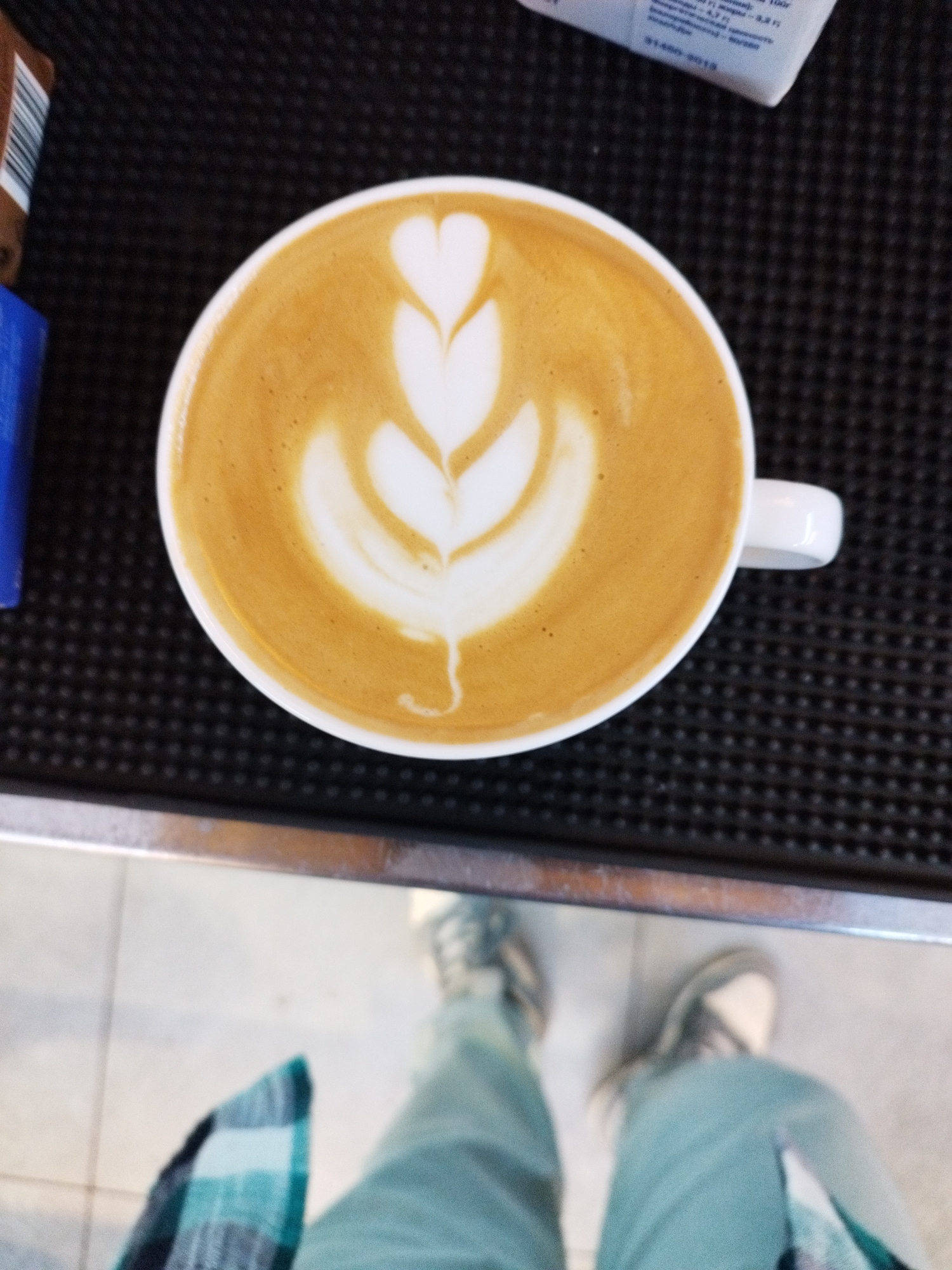 An overhead shot captures a latte with intricate foam art, resembling a tulip or swan, set against a dark textured surface. The coffee cup is white and sits on a black, textured mat. To the left of the cup, a small portion of a blue object and a brown and white striped object are visible. In the background, the lower legs and feet of a person wearing light blue pants and a checkered shirt are visible. The scene appears to be indoors, possibly in a cafe or a home kitchen. The specific location in Saint Petersburg, Russia cannot be determined from the image.
