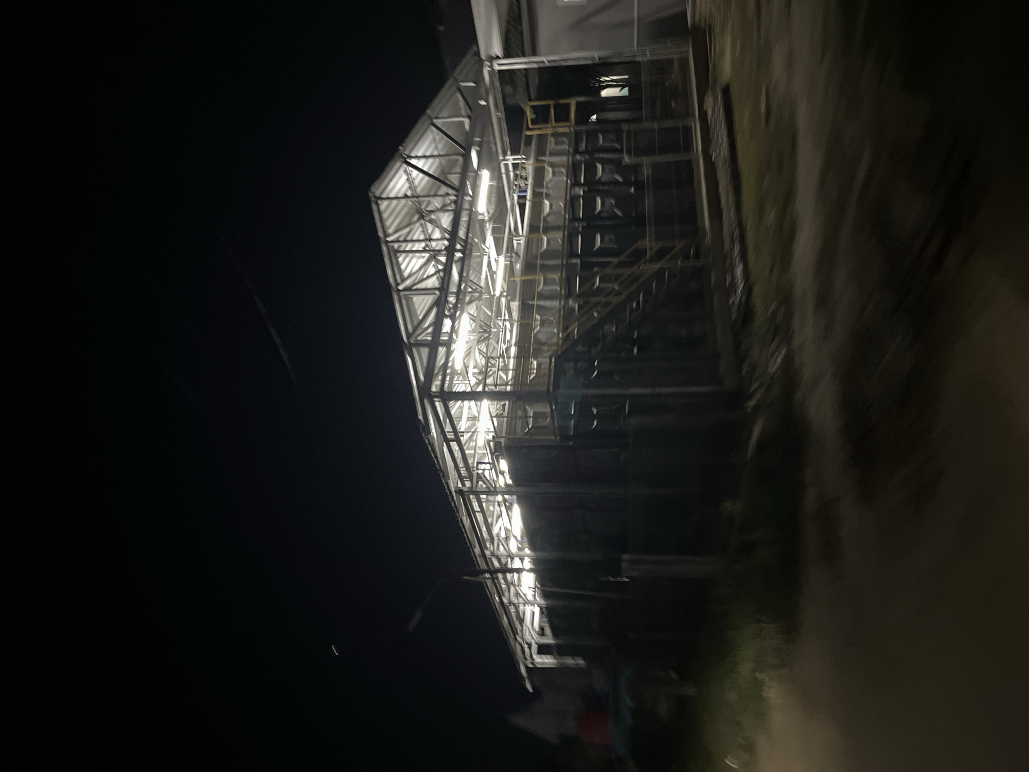At night, an illuminated, open-sided building with a metal frame and corrugated metal roof is visible. Inside, large, dark tanks are arranged in rows, with yellow metal stairs providing access to a raised platform. A street lamp stands in front of the structure. The foreground is blurred, suggesting movement, possibly from a vehicle. The background is dark, with a faint light from a distant building on the left and a crescent moon or thin cloud in the upper left sky. The image is taken at a location in the city of Kesehatanbandar, Indonesia.