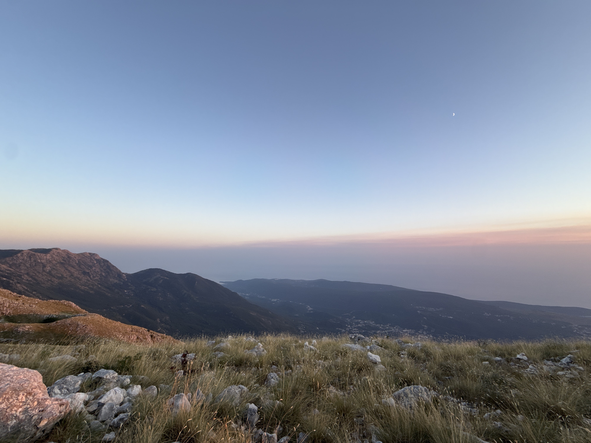 This image depicts a panoramic view from a grassy, rocky mountainside, likely in or near Kuk, Montenegro. The foreground is dominated by dry, wispy grass and scattered grey and pinkish rocks. In the mid-ground, rolling hills of varying shades of green and brown are visible, with some areas illuminated by soft, warm light, suggesting it is either dawn or dusk. Further in the distance, a hazy coastline stretches across the horizon, with a body of water, possibly the Adriatic Sea, meeting the sky. The sky itself is a gradient of clear blue at the top, transitioning to soft pinks and oranges closer to the horizon, indicating a sunrise or sunset. A faint crescent moon is visible in the upper right quadrant of the sky. There are no people or animals in the image. The overall scene is serene and natural, highlighting the vastness of the landscape.