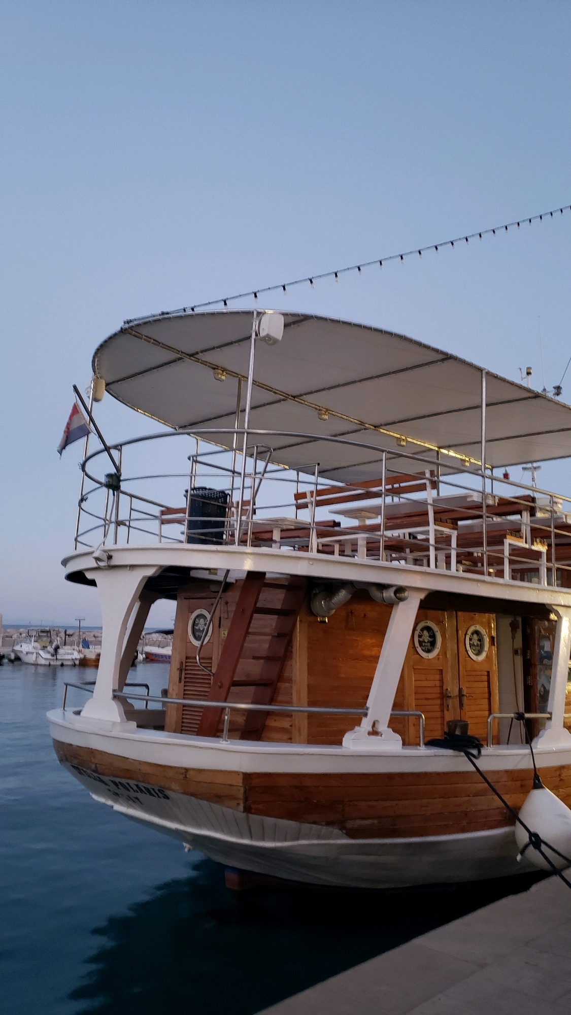 A wooden boat with a white canopy is docked in a harbor under a clear, pale sky. The boat has multiple levels with wooden benches and tables. A Croatian flag flies from the bow. The boat's name, "VENERA POLARIS," is visible on its hull. In the background, other boats are moored in the harbor. The water is a deep blue. The scene appears to be in Općina Tučepi, Croatia, a coastal municipality.