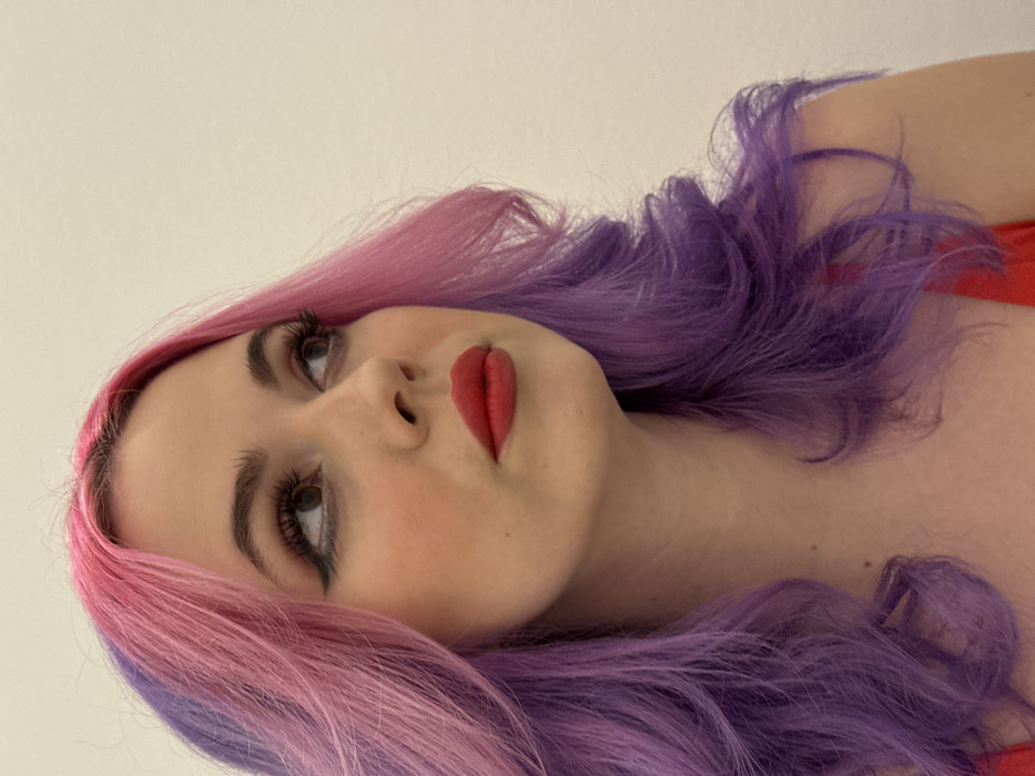 A woman with pink and purple hair is pictured with her head tilted toward the viewer. She has fair skin, full red lips, and dark eyelashes. Her gaze is directed upward and slightly to the left. A red garment is visible near her shoulder and neckline. The background is a plain, light color, possibly off-white.