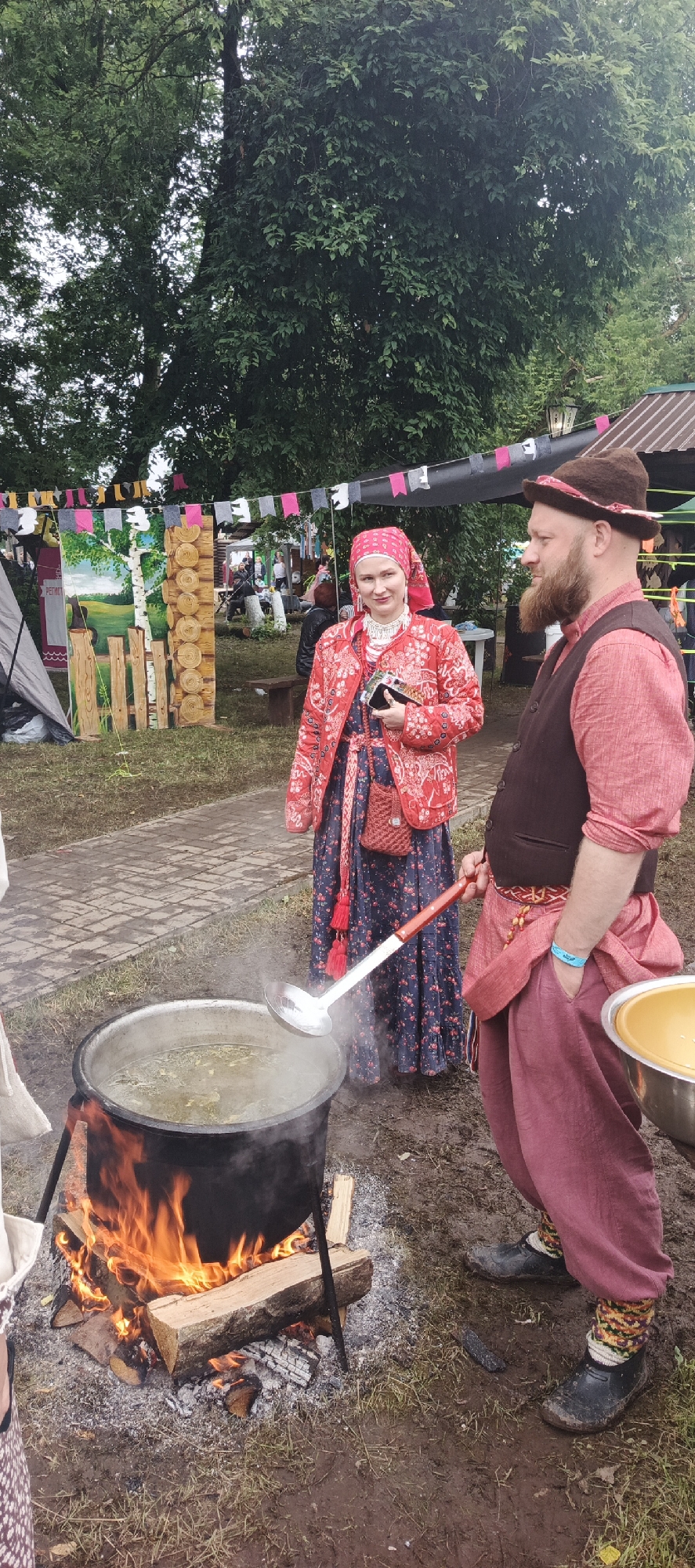 The image depicts an outdoor scene, possibly a festival or market, in the village of Старые Быги, Russia. In the foreground, a man and a woman are standing near a large pot of food being cooked over an open fire. The man, wearing a dark vest, a red shirt, and dark trousers, has a beard and a hat. He is looking to the right. The woman, wearing a traditional red embroidered jacket, a floral dress, and a red headscarf, is holding a ladle over the pot, which is steaming. Behind them, there are tents, decorative wooden structures, and trees, suggesting a rural or park-like setting. The ground is a mix of dirt and paved walkway. The overall atmosphere appears festive.