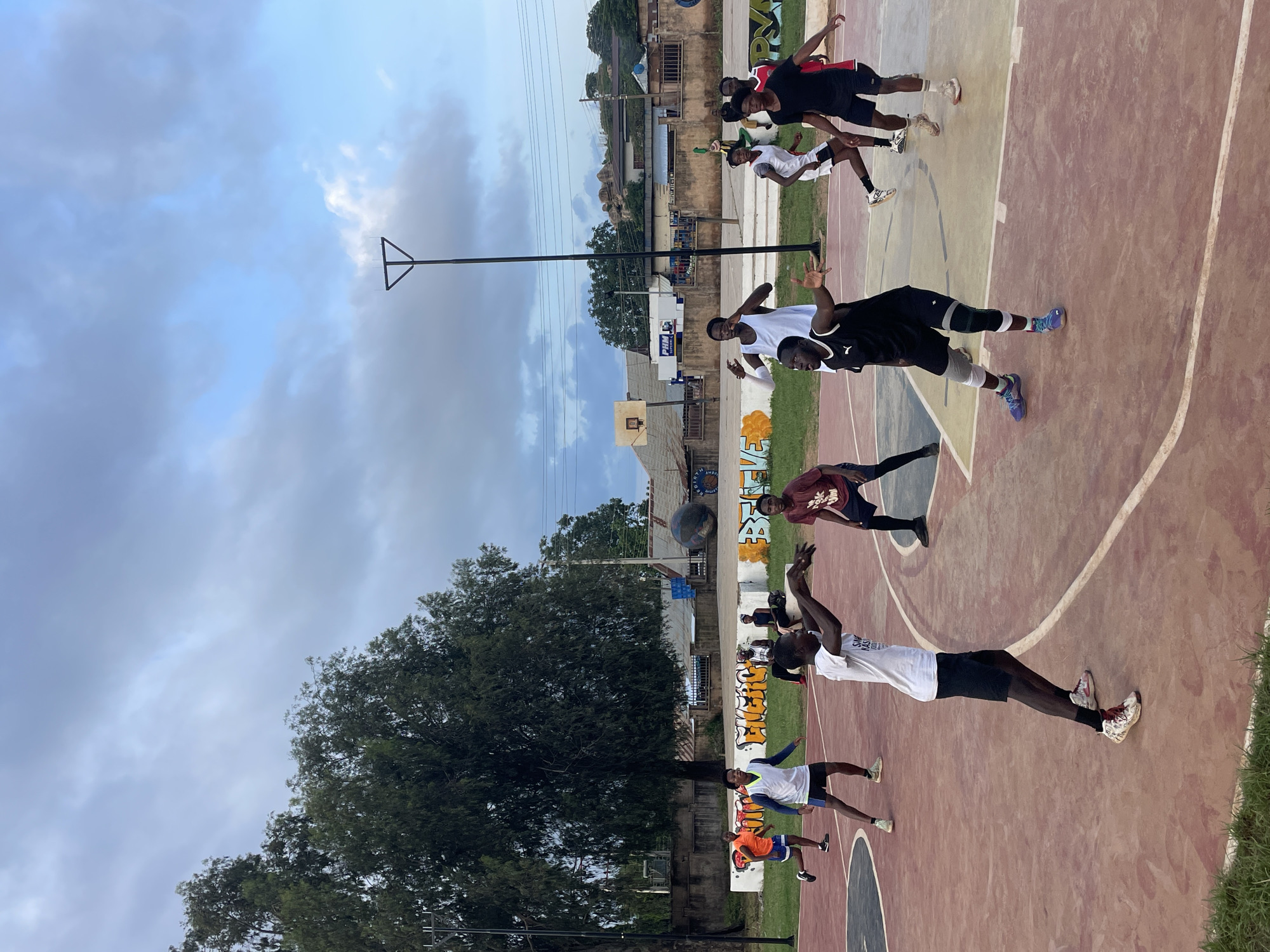 The image shows a group of young people playing basketball on an outdoor court in Gura Top, Nigeria. The sky is cloudy, suggesting it might be late afternoon or early evening. The court itself is a mix of red and yellow surfaces, with white lines marking its boundaries. The players are dressed in casual athletic wear, and some are actively engaged in the game, while others appear to be waiting their turn.
Notable details include a basketball hoop and backboard, a basketball in the air, and graffiti on a nearby wall that says "SMASH KINGS." The surrounding area appears to be a mix of urban and natural elements, with buildings, trees, and power lines visible in the background. The overall atmosphere seems energetic and youthful.