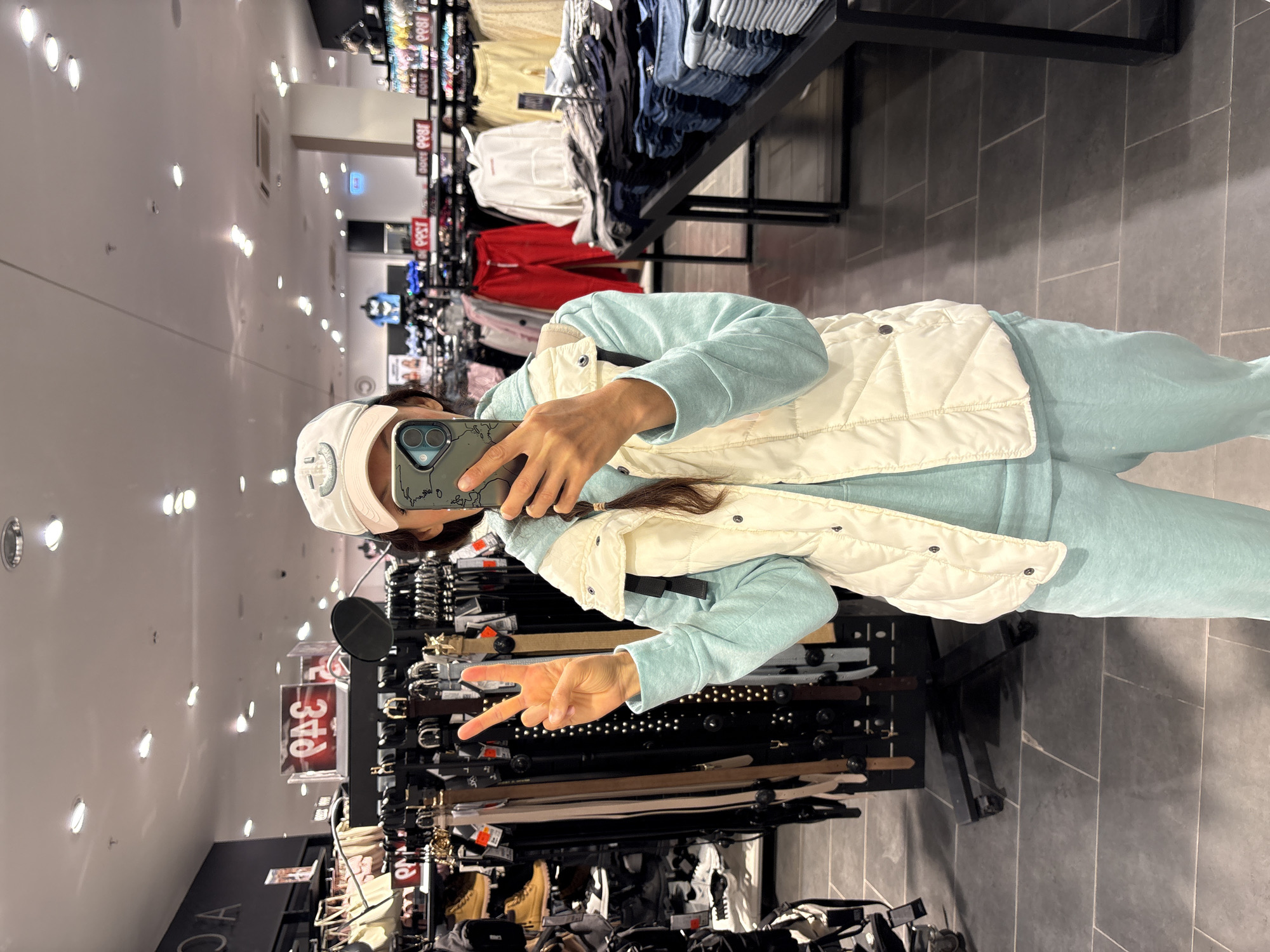 A person stands in a retail store in Ulyanovsk, Russia, taking a selfie. They are wearing a light blue sweatsuit and a white quilted vest, with a white baseball cap on their head. Their hair is pulled back in a braid. The person is holding a phone with a green case, using it to capture the image. They are making a peace sign with their left hand.
In the background, shelves display various items, including belts and other clothing. Overhead lighting is visible in the ceiling. The floor is tiled in gray. Clothes are organized on shelves and racks, indicating a fashion or clothing department within the store.