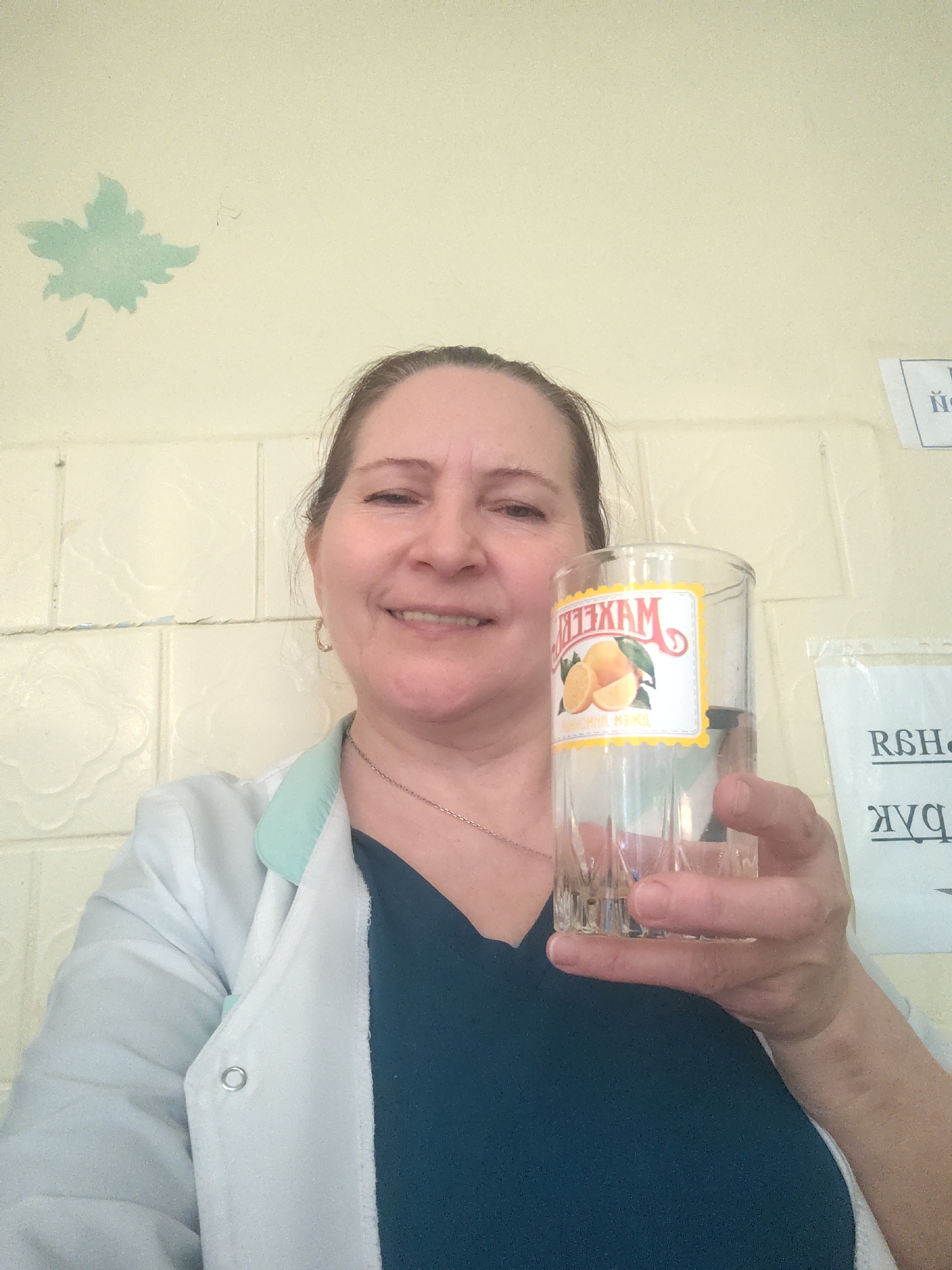 A smiling, fair-skinned woman, likely in her 40s or 50s, is captured in a close-up selfie. Her light brown hair is pulled back, and she wears a thin silver necklace. She is dressed in a white lab coat with a light blue collar trim over a dark blue top.
The woman is holding a clear glass tumbler in her right hand, presenting it towards the camera. The glass features a rectangular label with a yellow border. On the label, the brand name "MAXELL" (МАХЕЛЛ) is prominently displayed in Cyrillic, along with an illustration of oranges and the phrase "апельсиновый макс" (orange max), indicating an orange-flavored product.
The background consists of a light cream-colored wall. On the left side of the upper wall, there is a stenciled or stickered light teal, five-lobed leaf design. The lower part of the wall features textured, off-white tiles arranged in a repetitive, ornate pattern. To the right of the woman, a white paper sign with partially visible, reversed black Cyrillic text is taped to the wall.
The woman is smiling broadly, conveying a cheerful and friendly demeanor. The scene appears to be indoors, under typical artificial or diffused natural light. The visible Cyrillic text on the glass and the background sign are consistent with the provided location context of Sarapul, Russia.