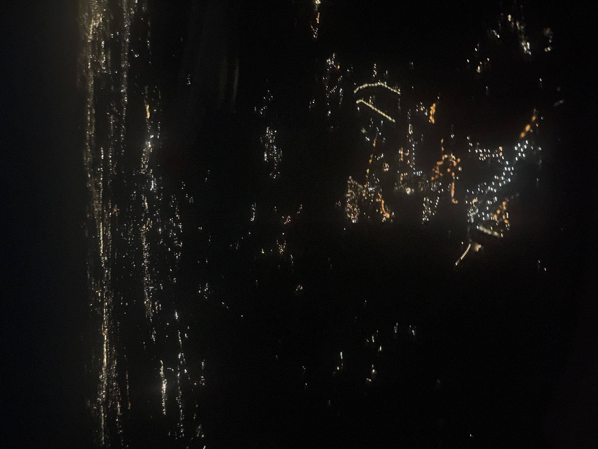 The image shows an aerial view of a city at night. The city's lights are visible as bright spots and streaks against the dark background. There is a dense concentration of lights on the right side of the image, which thins as it spreads to the left. Due to the darkness and aerial perspective, specific details about the location and city of Trevignano Romano, Italy, are unidentifiable. No people, specific subjects, objects, actions, or interactions are visible. The environment is characterized by a city illuminated at night.