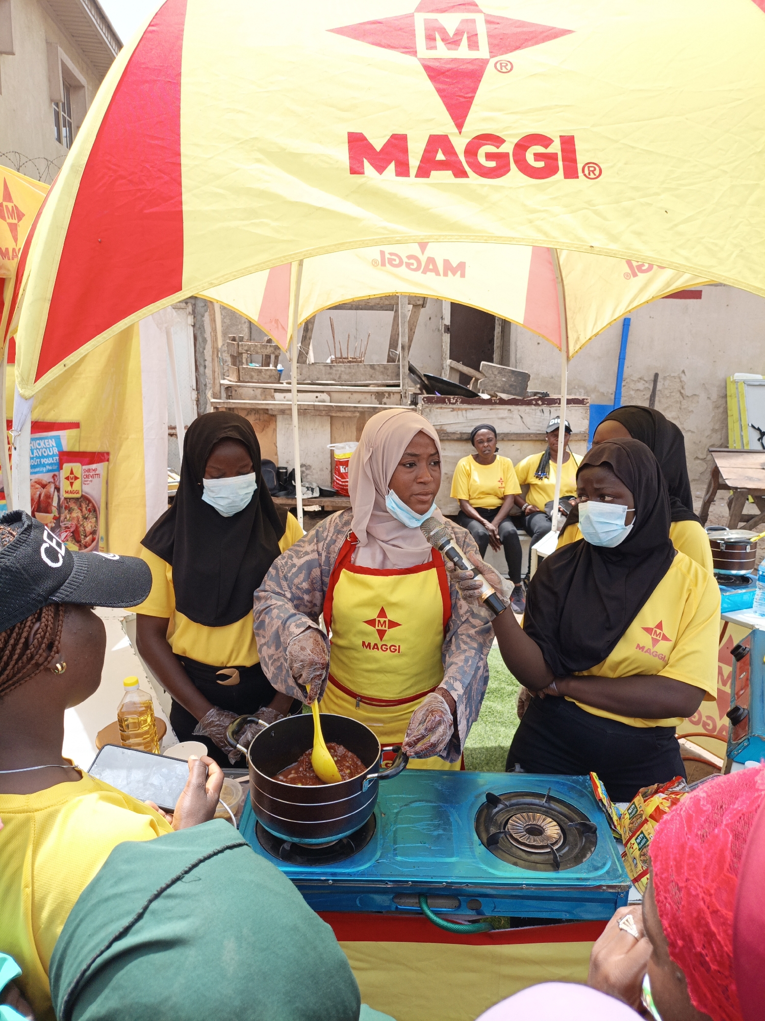 A medium shot captures an outdoor cooking demonstration event, likely in Tarauni, Nigeria, under a red and yellow branded canopy displaying "MAGGI." In the foreground, three young women are the primary subjects. On the left, a woman wearing a yellow shirt and a black cap with braided hair and gold earrings is partially visible, holding a smartphone. In the center, a woman in a black hijab and a yellow shirt, wearing a light blue surgical mask, is looking down at a pot on a blue gas stove. She is stirring a reddish sauce with a yellow spoon, wearing clear plastic gloves. To her right, a woman in a light brown hijab and a yellow apron over a patterned long-sleeved shirt, also wearing a blue surgical mask, is speaking into a microphone held by the central figure. In the background, other individuals, some wearing yellow shirts with the Maggi logo, are seated or standing. The environment appears to be an urban setting with buildings visible behind the canopy and some construction materials in the background. Several Maggi product packages are displayed on shelves behind the cooking area. The overall scene depicts a promotional event or a cooking class focused on Maggi products.