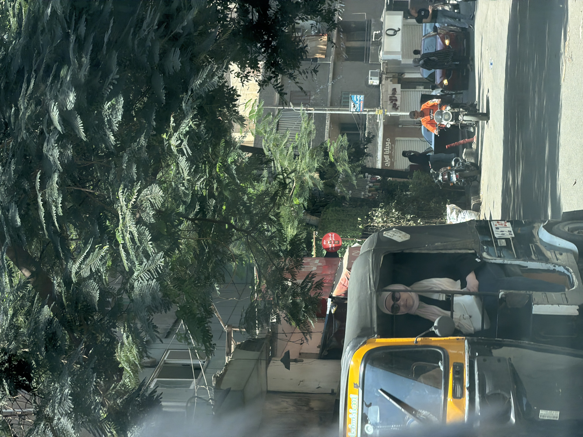 In Cairo, Egypt, on a sunny day, a street scene unfolds with a bright yellow and black auto-rickshaw in the foreground. A woman wearing a light-colored hijab and sunglasses sits in the passenger seat of the auto-rickshaw, holding a light-colored bag. The auto-rickshaw is stopped or moving slowly, as indicated by the woman's relaxed posture. Behind the auto-rickshaw, a man in an orange shirt rides a motorcycle, looking straight ahead. Cars and other pedestrians are visible further down the street. The buildings lining the street appear to be old, with visible signs of wear and tear, including peeling paint and exposed wiring. Lush green trees with dense foliage provide shade over the street, creating a dappled sunlight effect. The overall atmosphere suggests a typical urban environment in Cairo, with the hustle and bustle of daily life captured in this moment.