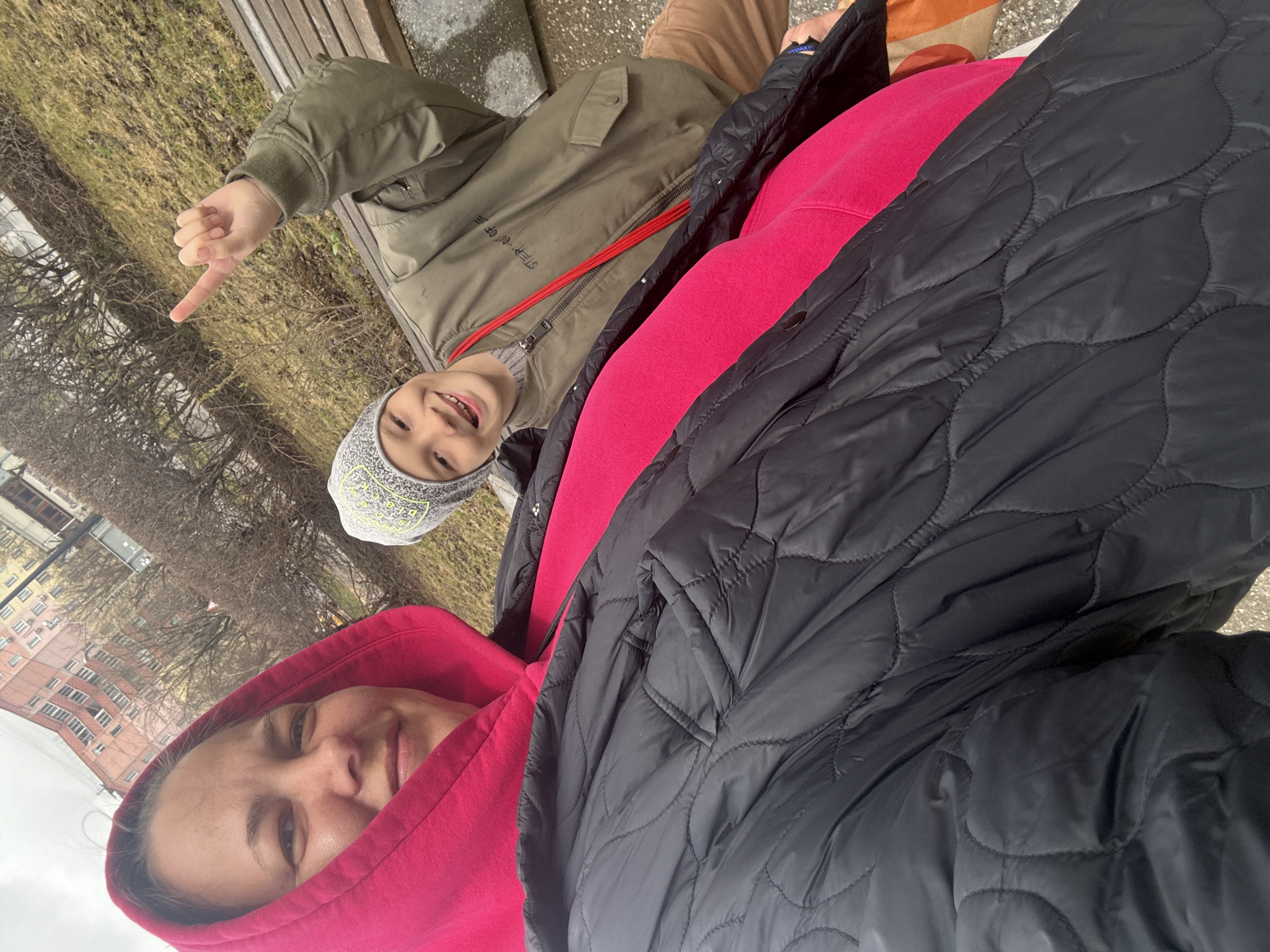 The image is a selfie of a woman and a child, possibly a mother and son, taken outdoors in Dmitrov, Russia. The woman is wearing a pink hoodie and a black quilted jacket. Her face is visible in the lower left corner. The child is wearing a gray beanie and a green jacket with a red strap. The child is smiling and pointing to the left. They are sitting on a wooden bench in front of bare trees. In the background, there are buildings typical of Dmitrov, including a light-colored building with many windows. The ground is paved and covered with some debris.