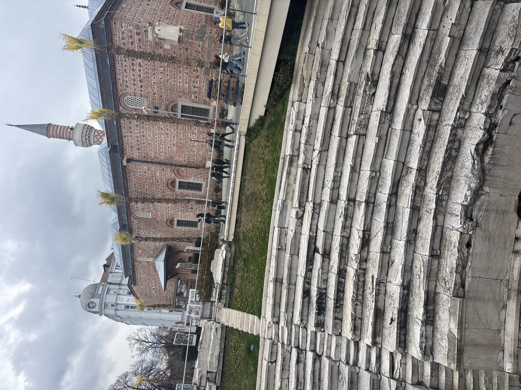 The image depicts a historical urban scene in Plovdiv, Bulgaria, focusing on the Roman Stadium complex. In the foreground, a significant section of ancient, light-grey stone amphitheater seating dominates the lower half of the frame, showing weathered, horizontally stacked tiers with visible cracks and dark discoloration.
Above the stadium seating, a grassy slope featuring scattered yellow flowers ascends to a paved area. A set of white stone steps cuts through this slope, connecting to a higher level. Several large, irregular white stone blocks are positioned on the grass.
Running across the mid-ground is a modern elevated pedestrian walkway with clear panels and metal railings. Approximately nine individuals are present on this walkway. A group of three to four people walks from left to center, and a second group of four to five people, including one individual in a yellow jacket, walks further right. A rectangular planter box filled with purple and orange flowers sits on the walkway's right side.
In the background, directly behind the walkway, stands the Dzhumaya Mosque, characterized by its reddish-brown brickwork with patterned lighter accents and a multi-segmented blue-grey roof. A tall, striped minaret with a conical spire rises from its rear-right. Several slender palm trees are positioned along the mosque's facade. To the left of the mosque, a multi-story, light-colored building with a domed clock tower is visible. Bare deciduous trees are present in the far left background. The sky is uniformly overcast with grey clouds.
