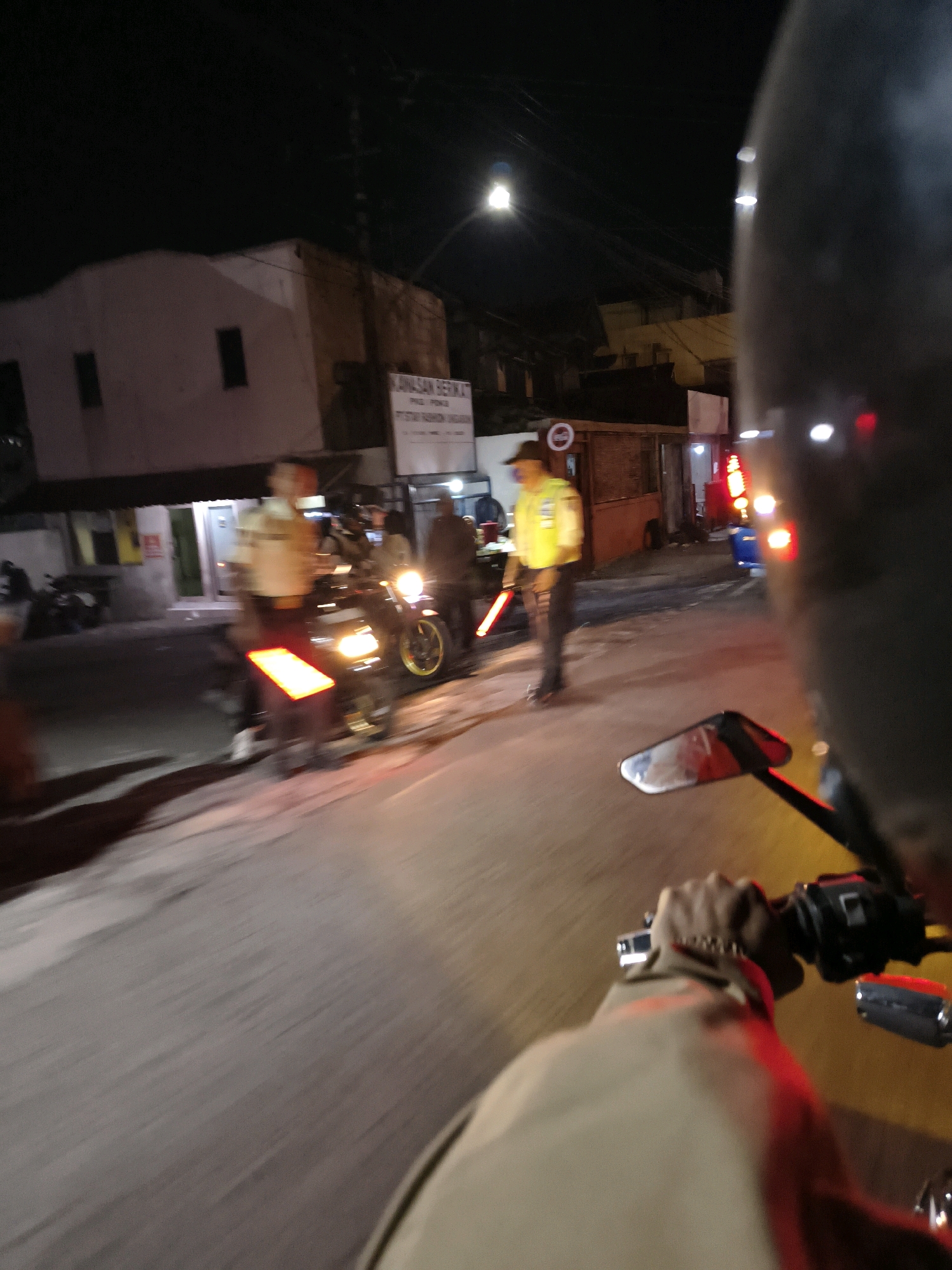 The image depicts a nighttime scene on a street in Bawen, Indonesia, likely near a police checkpoint or traffic stop. In the foreground, a person's hand is visible, gripping a motorcycle handlebar. The rider's perspective suggests they are approaching or are part of the scene. In the middle ground, several individuals in uniform, appearing to be police officers, are present. One officer in a yellow vest and dark pants holds a glowing red traffic baton, directing traffic. Another officer is visible in the background. A motorcycle with its headlights on is positioned on the road, and another motorcycle is parked on the side. Buildings line the street, some with illuminated windows. The overall atmosphere is one of activity and order enforcement on a dark street.