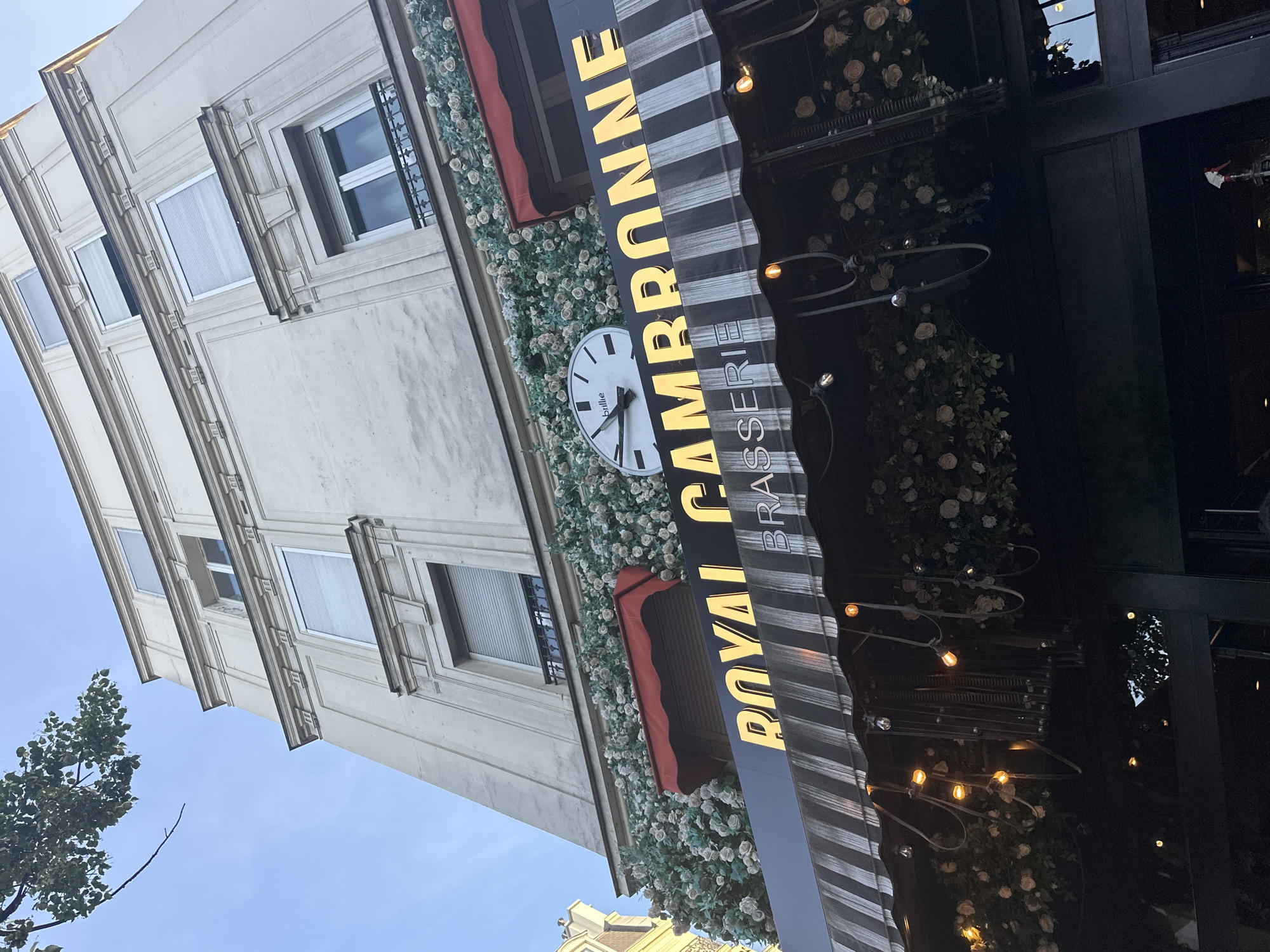 The image captures a low-angle view of the "ROYAL CAMBRONNE BRASSERIE" located in Paris, France. The brasserie features a prominent awning with black and white stripes, adorned with decorative string lights and artificial white roses. Above the awning, the facade of a building is visible, with several windows and ornate architectural details. A clock with a white face and black hands is mounted on the wall above the brasserie's signage. The sky is a clear blue with a few green leaves from a tree visible in the upper left corner. The overall scene suggests an outdoor dining or establishment setting in Paris.