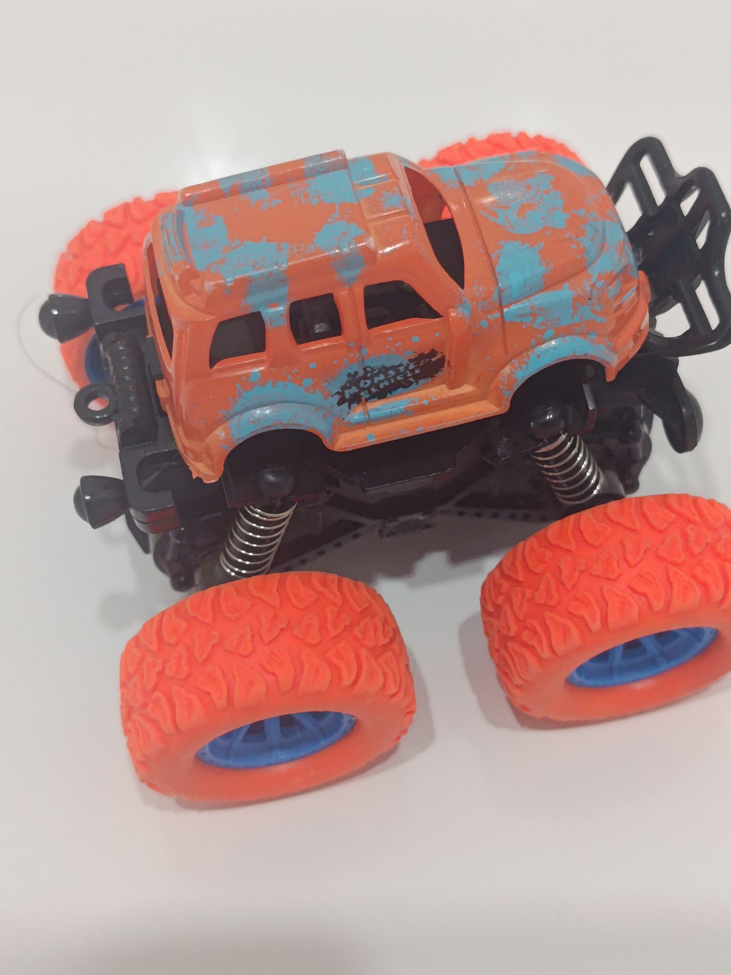 The image shows a toy monster truck, primarily orange with distressed blue paint. The truck has large, knobby orange tires with blue rims. The chassis and suspension are black, with visible metal springs. The toy is positioned on a white surface, likely a table or floor, and is seen from an elevated, slightly angled perspective. The background is entirely white, offering no context for its location. The toy is the sole subject in the frame.