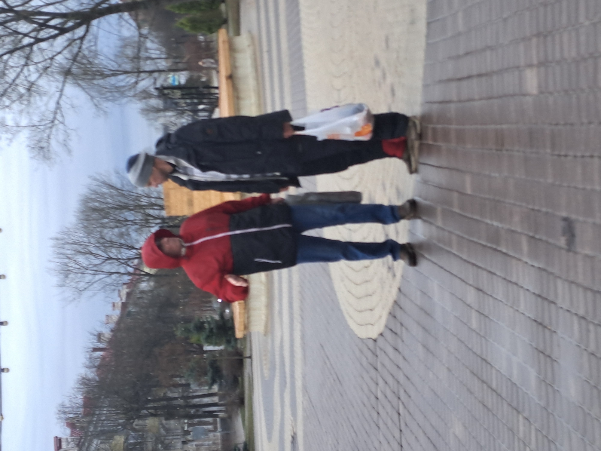 A photograph depicts two people walking on a brick-paved surface. The individual closest to the foreground is wearing a red hooded jacket, blue jeans, and dark shoes. The second person is behind them, wearing a black jacket, grey hat, and dark shoes, and is carrying a plastic bag. Both are walking in the same direction. The background features bare trees suggesting a fall or winter season. In the distance, building structures and a park can be seen. The location is Elektrostal, Russia.