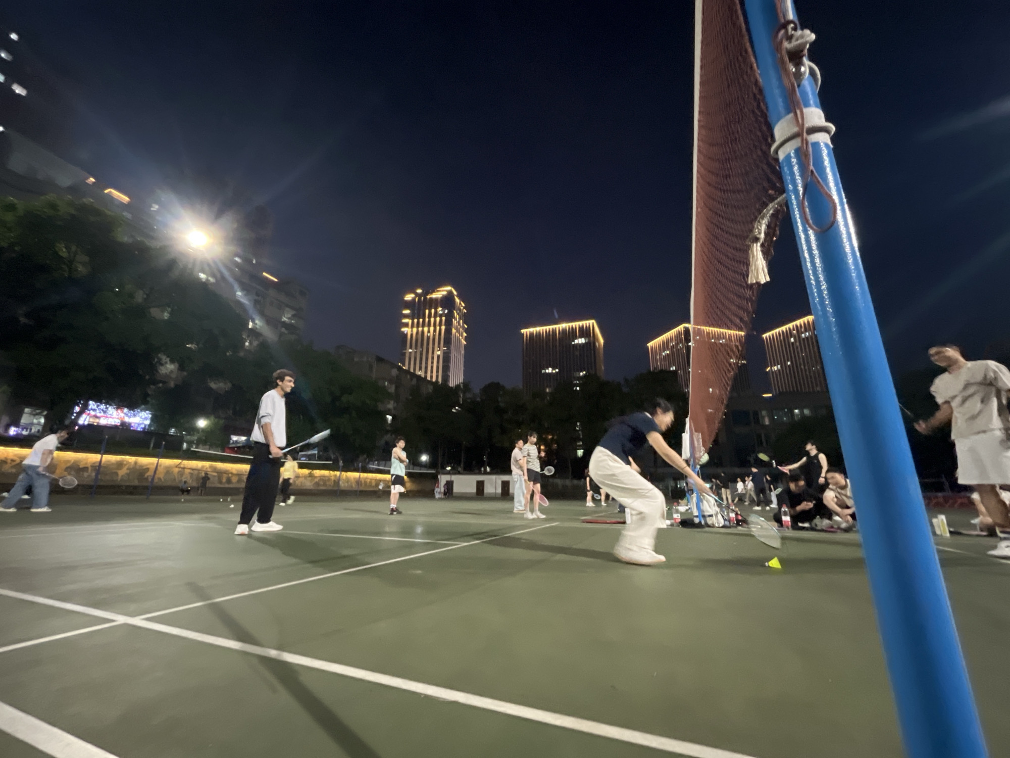 In Yuzhong District, China, at night, a group of people are playing badminton on an outdoor court. The court is green with white lines, and a blue pole supporting a red net is prominently visible in the foreground. Several individuals are actively engaged in the game, holding badminton rackets and positioned on the court. Others are observing the game or gathered nearby. In the background, illuminated by artificial lights, are tall buildings with distinctive gold lighting, suggesting an urban environment. The sky is dark, indicating it is nighttime, and trees are visible along the perimeter of the court. The scene captures a lively atmosphere of recreation in a city setting after dark.