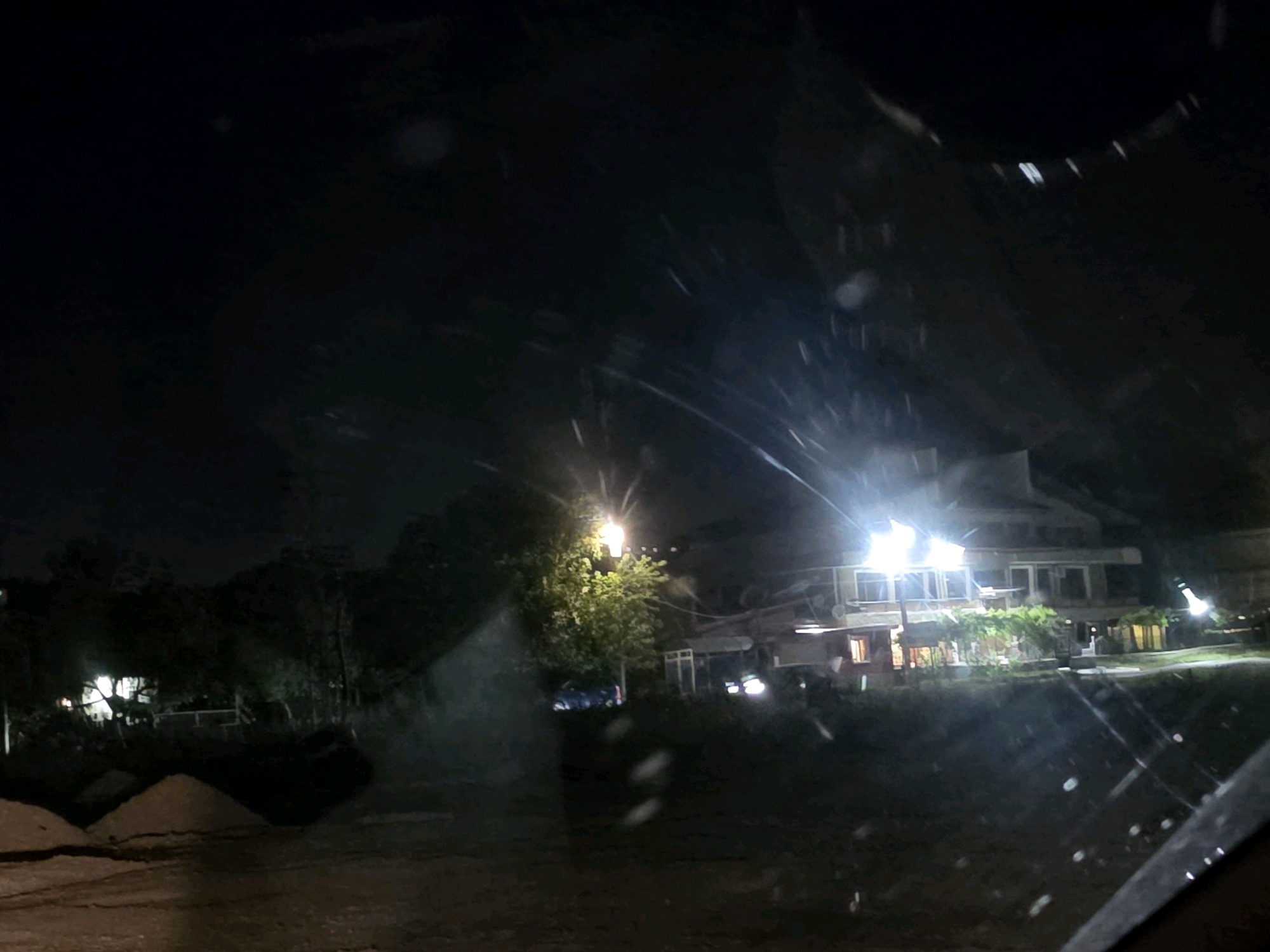 This is a nighttime photograph taken from what appears to be inside a vehicle, looking out through a dirty or wet windshield. The scene depicts a building with multiple illuminated windows and exterior lights, set against a dark, tree-lined background. In the foreground, there are mounds of material, possibly sand or dirt, and the ground is wet and reflective, suggesting recent rain or puddles. The lighting is artificial and somewhat hazy, with streaks and flares of light indicating the low visibility conditions and potential atmospheric interference. The overall impression is one of a dimly lit, possibly rural or suburban area at night, with the building being the primary point of interest.